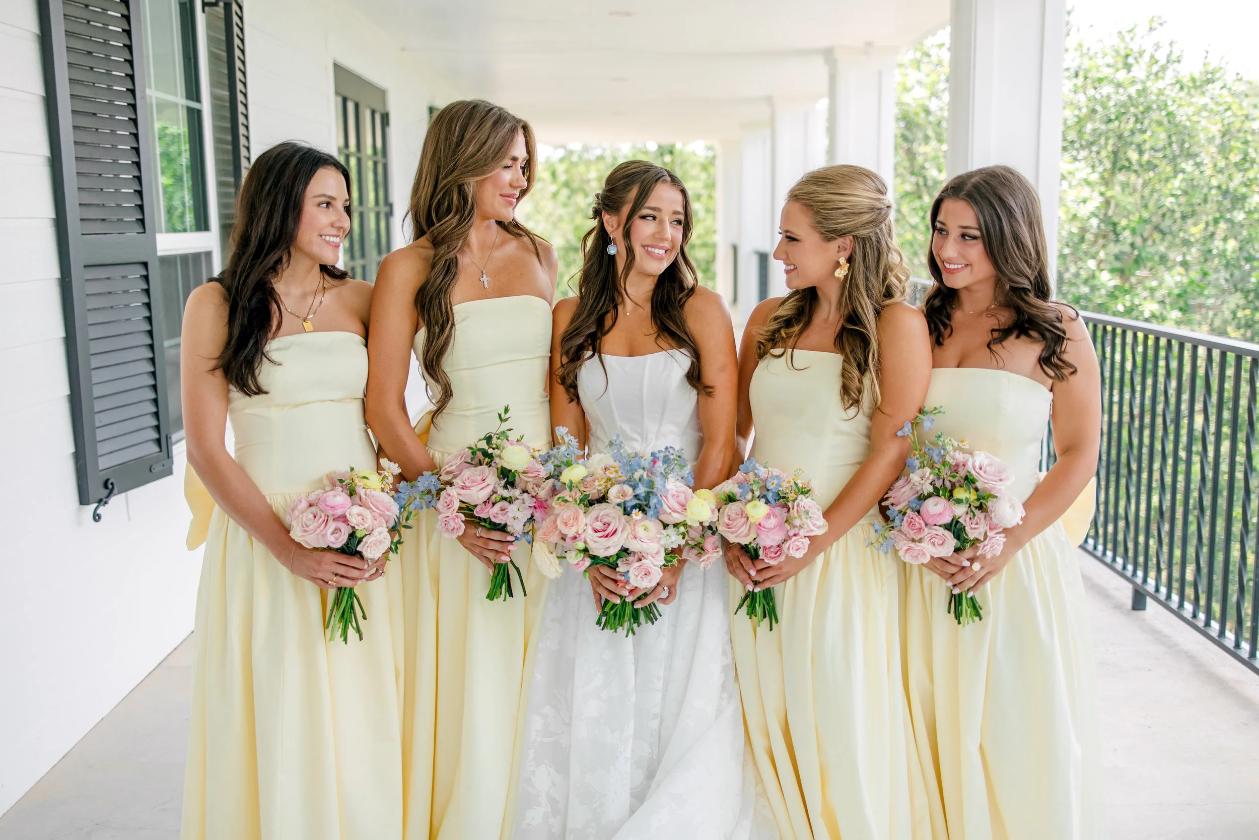 A bride and her five bridesmaids standing on a porch, all holding bouquets of pink and purple flowers, dressed in pastel yellow dresses.