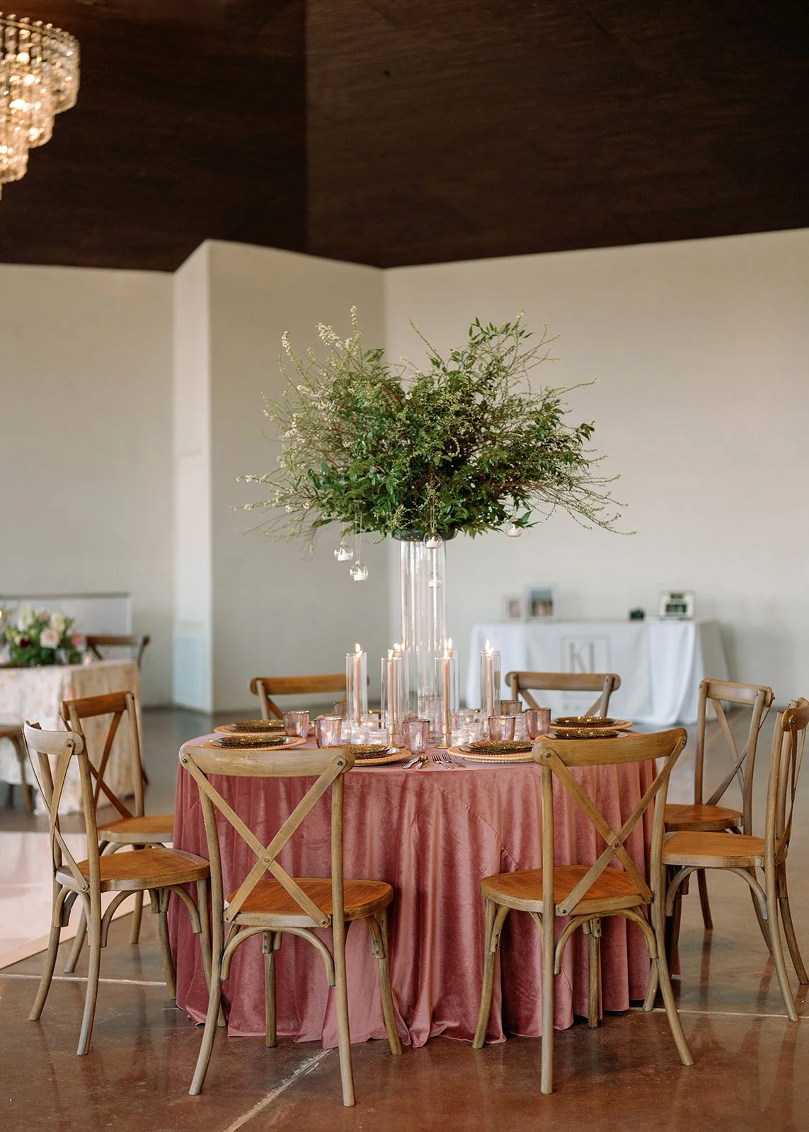 A round table with a pink tablecloth set for a formal event, featuring six place settings with gold chargers, pink glasses, and silverware. A tall, clear glass vase with a large arrangement of greenery and white flowers is at the center. The table is