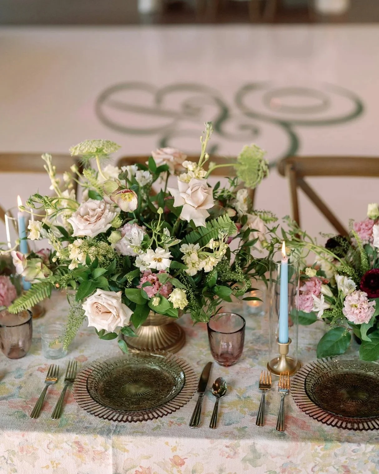 A table setting with a floral centerpiece, textured glass plates, silverware, pink and purple drinking glasses, a blue candle, and a pink cloth tablecloth.