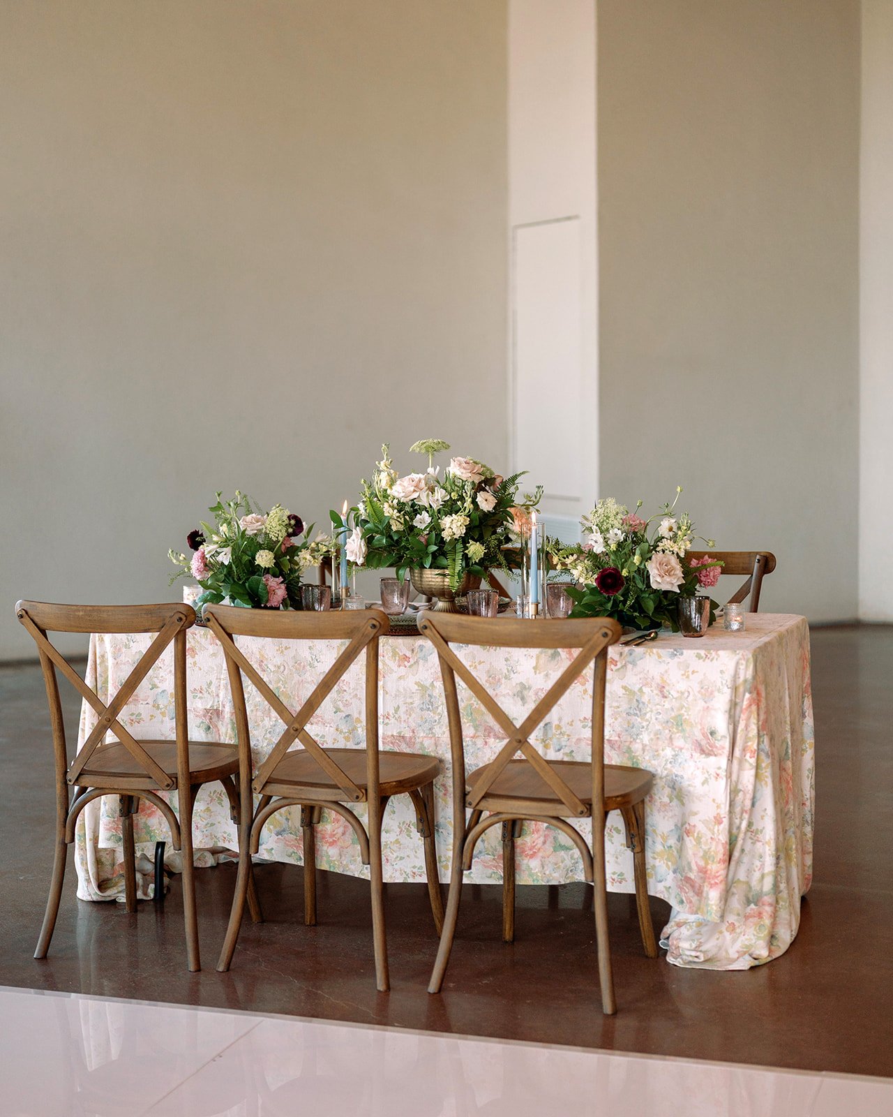 A rectangular table with a floral tablecloth, set with floral arrangements, candles, and glasses, surrounded by wooden chairs, in an empty room.