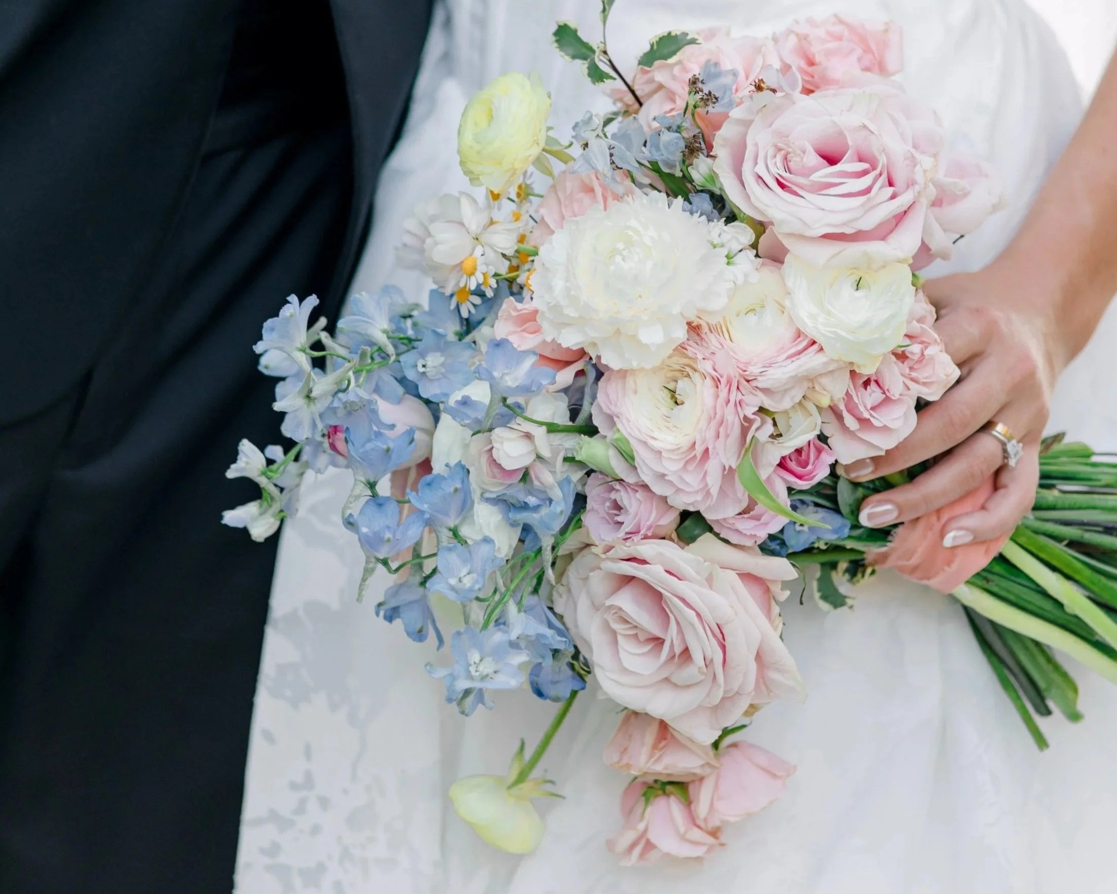 A bride holding a bouquet of pink, white, and light blue flowers, with a groom partially visible beside her.