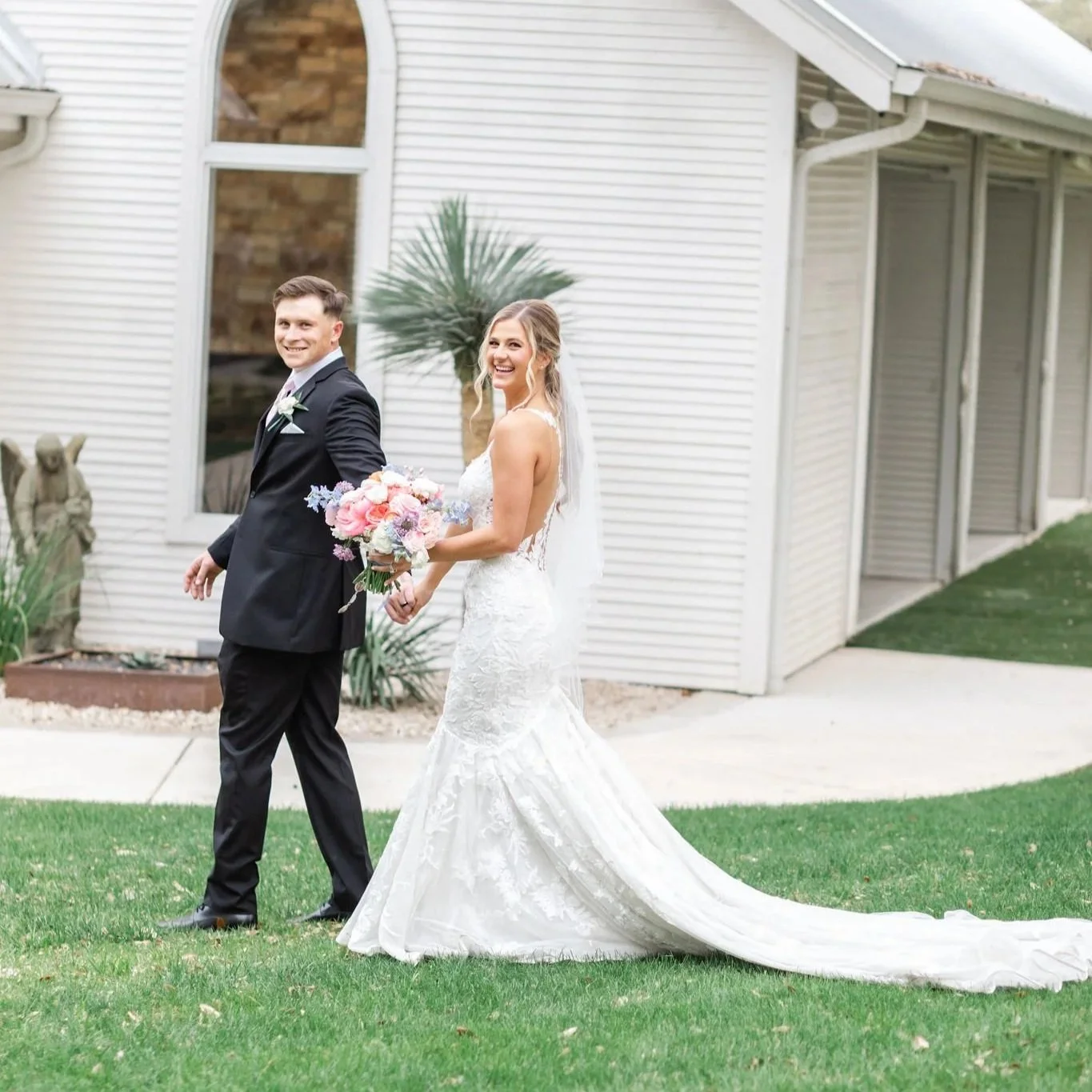 Bride and groom walking outside on their wedding day, smiling. The bride wears a white lace wedding gown with a train and veil, holding a bouquet of pink and purple flowers. The groom wears a black suit with a white shirt and black tie, smiling at the camera. They walk on green grass near a white house with large windows and a potted plant.