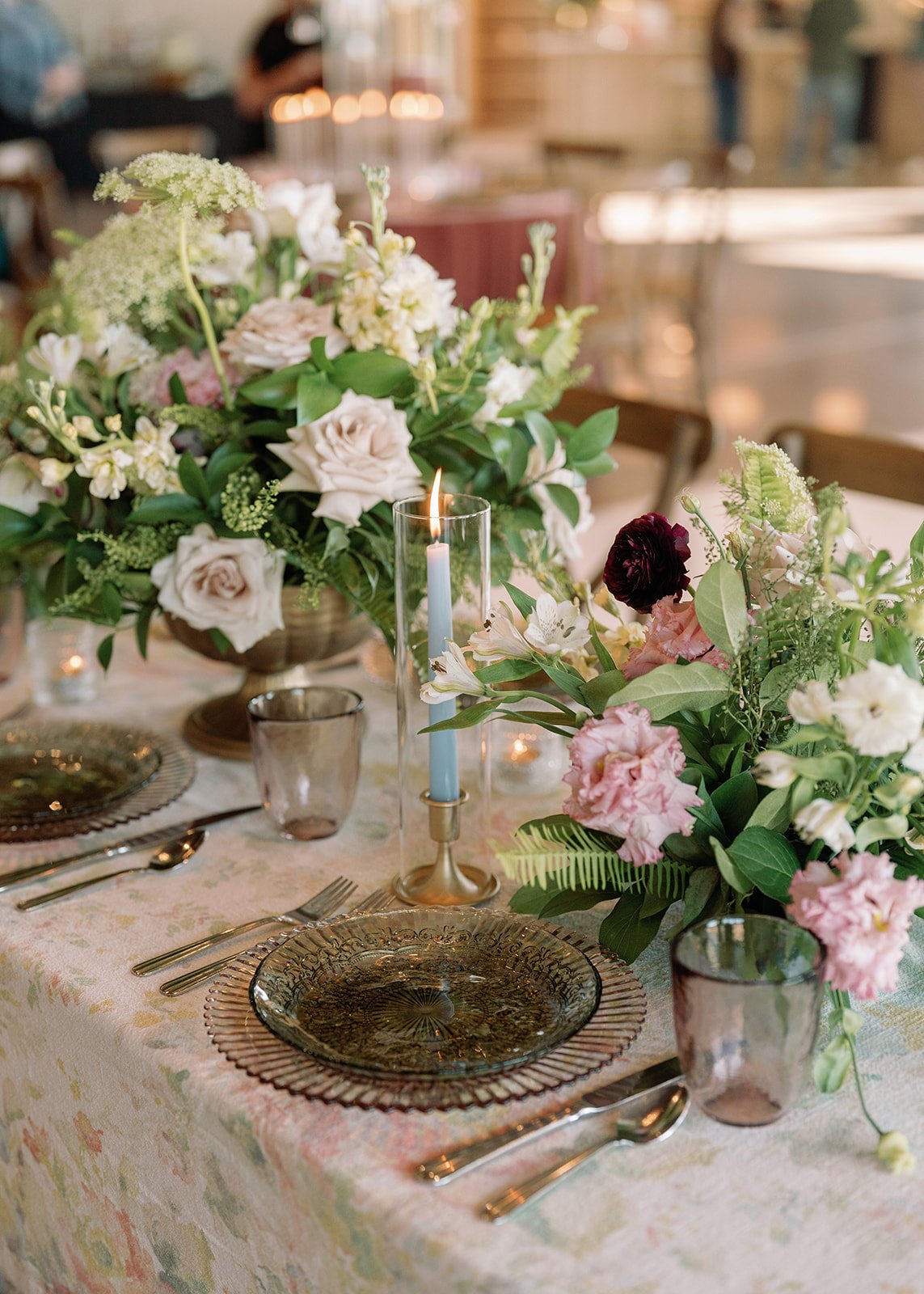 A wedding table setting with a floral centerpiece, glass plates, silverware, and a lit candle in a tall glass holder, in a decorated event space.
