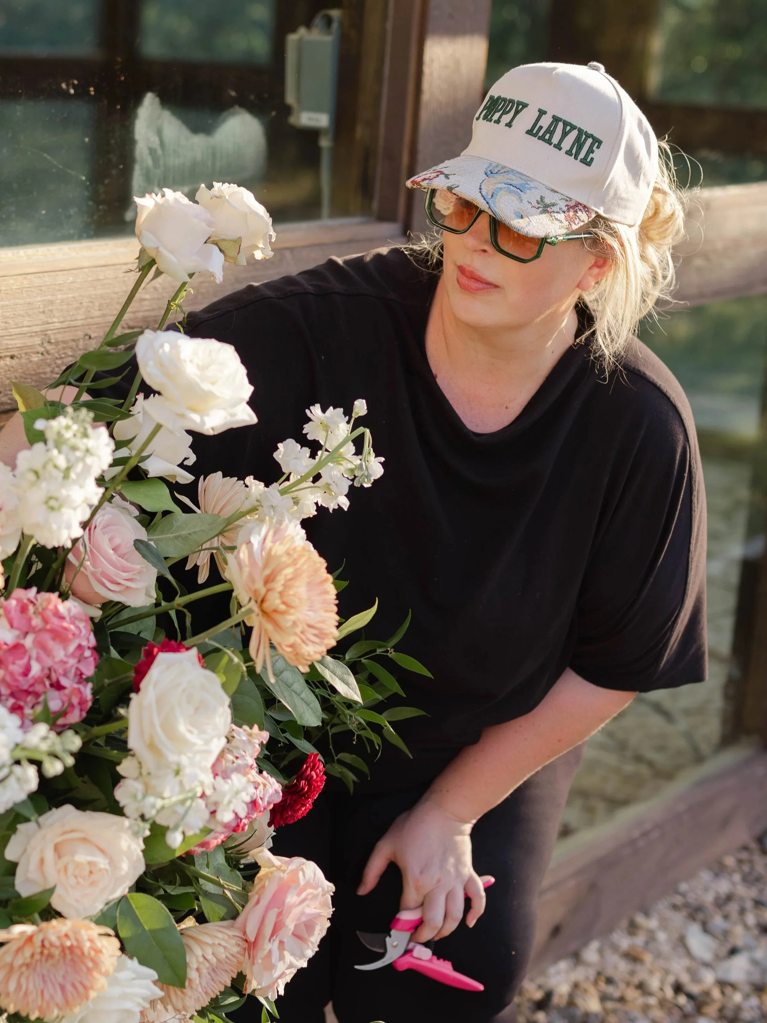 Woman wearing a white cap with green text that says 'Poppy Lane,' sunglasses, and a black shirt, tending to a colorful flower arrangement outside.