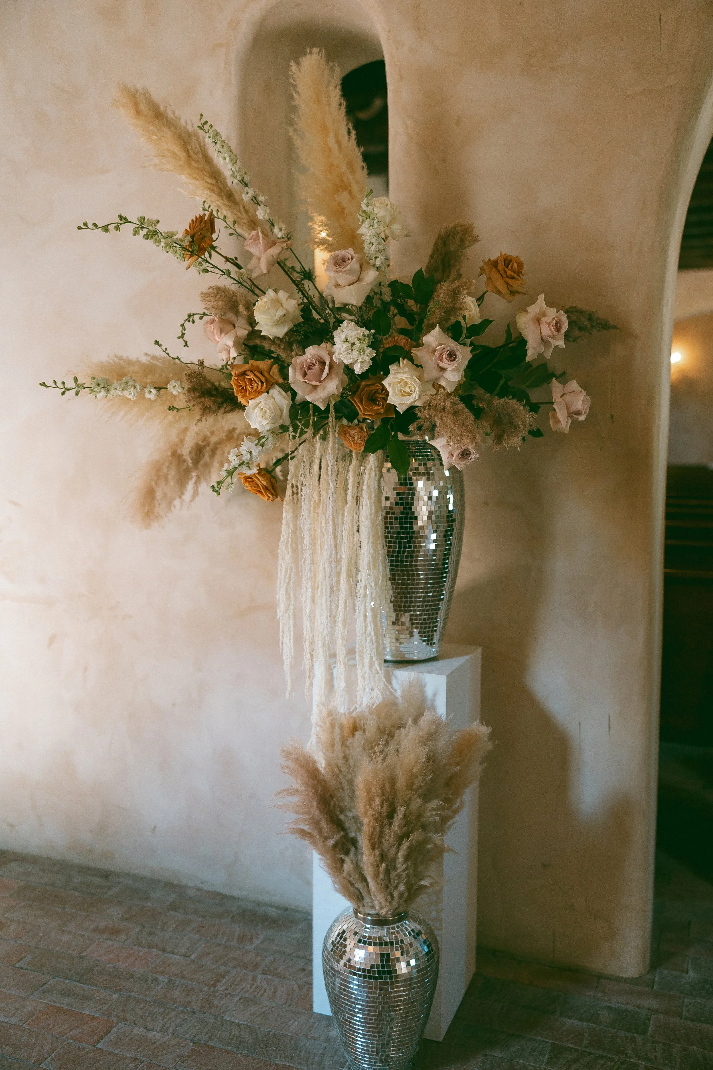 A large floral arrangement in a shiny, silver mosaic vase with beige, cream, white, and orange roses, pampas grass, and other flowers, placed on a white pedestal against a beige wall.