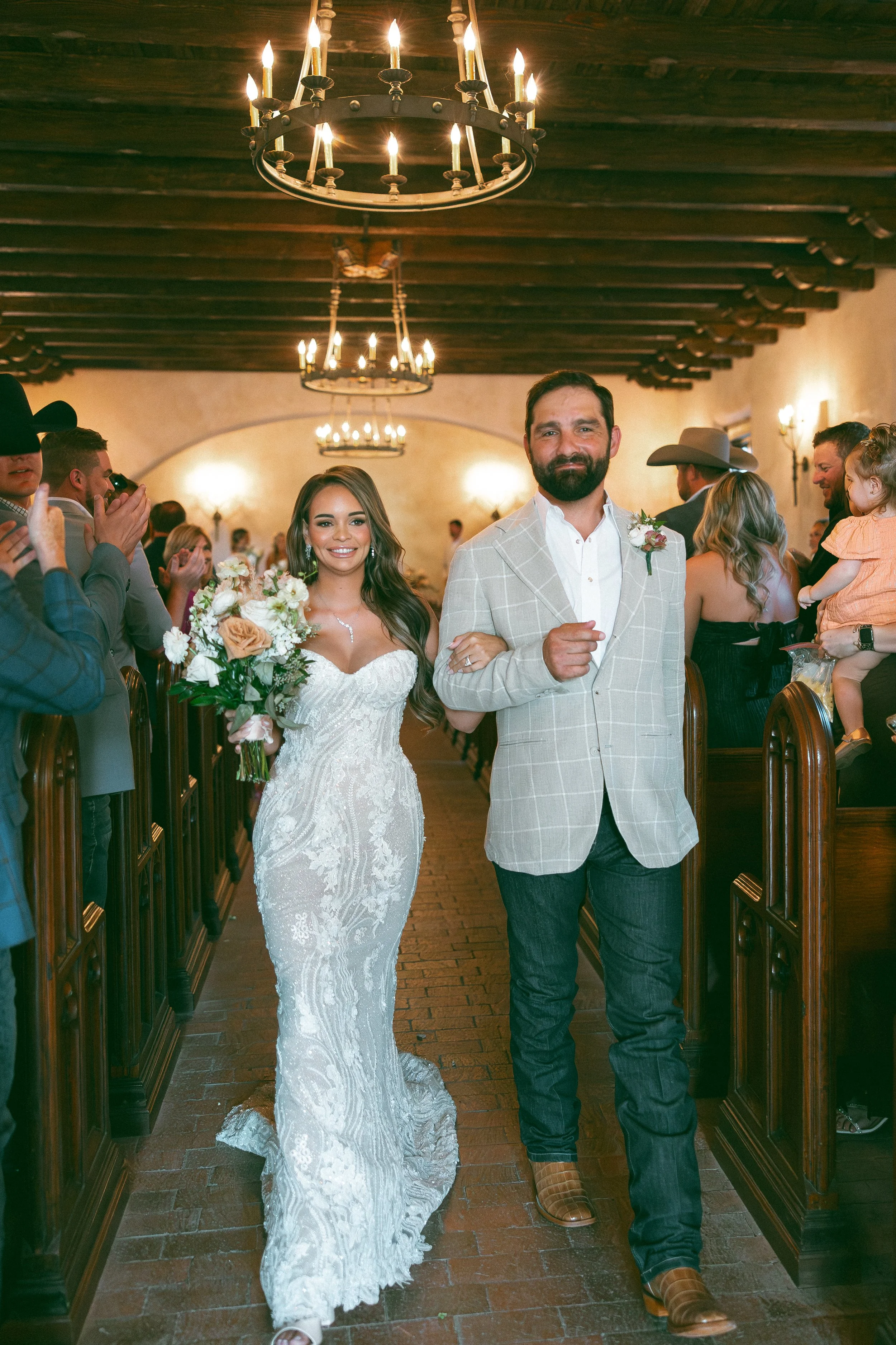 A bride and groom walking down the aisle in a wedding ceremony, surrounded by guests clapping and smiling inside a rustic wooden venue with chandeliers.