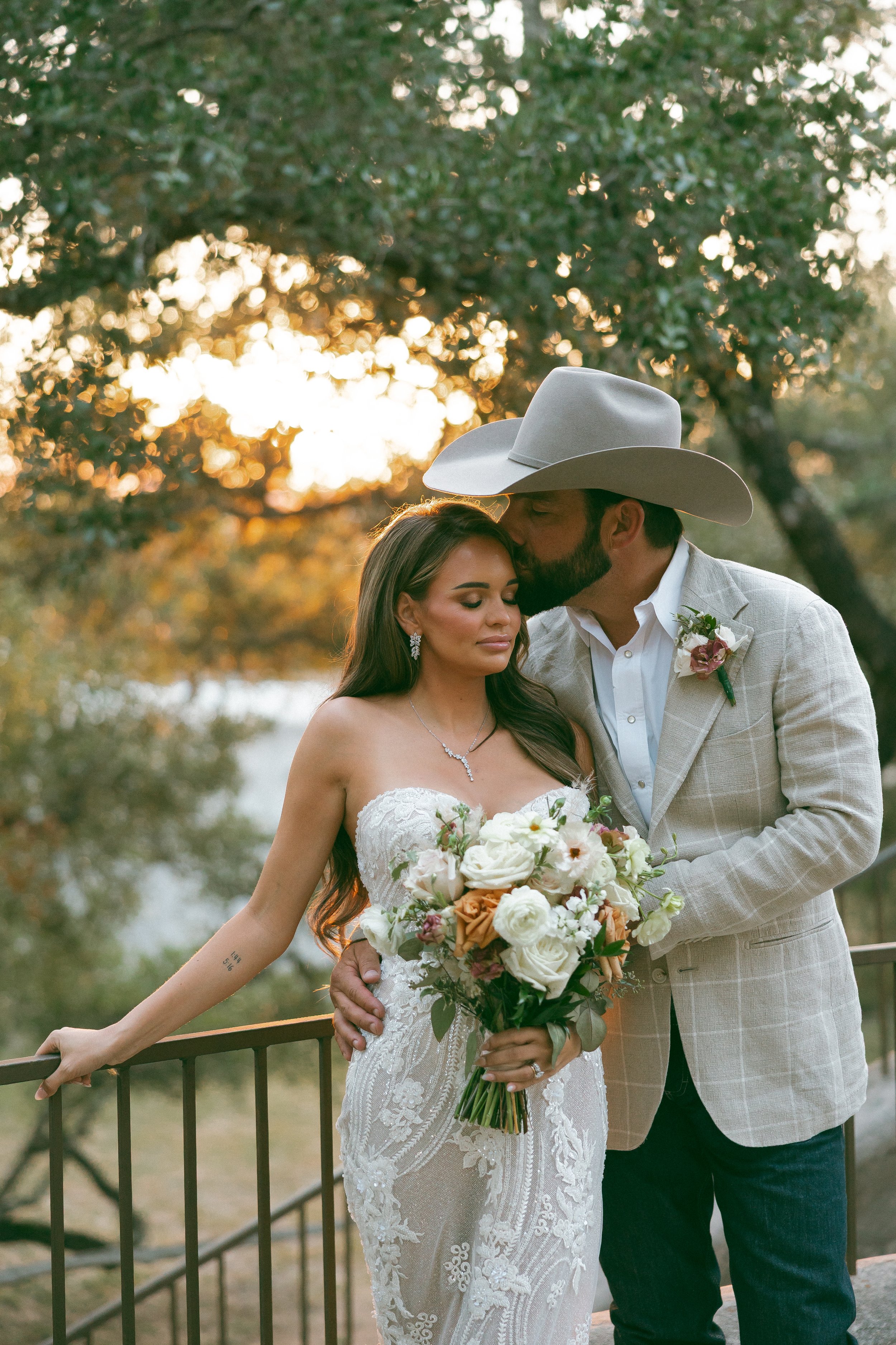 A bride and groom share a romantic moment outdoors during sunset, with the groom kissing the bride's forehead as she holds a bouquet of flowers.