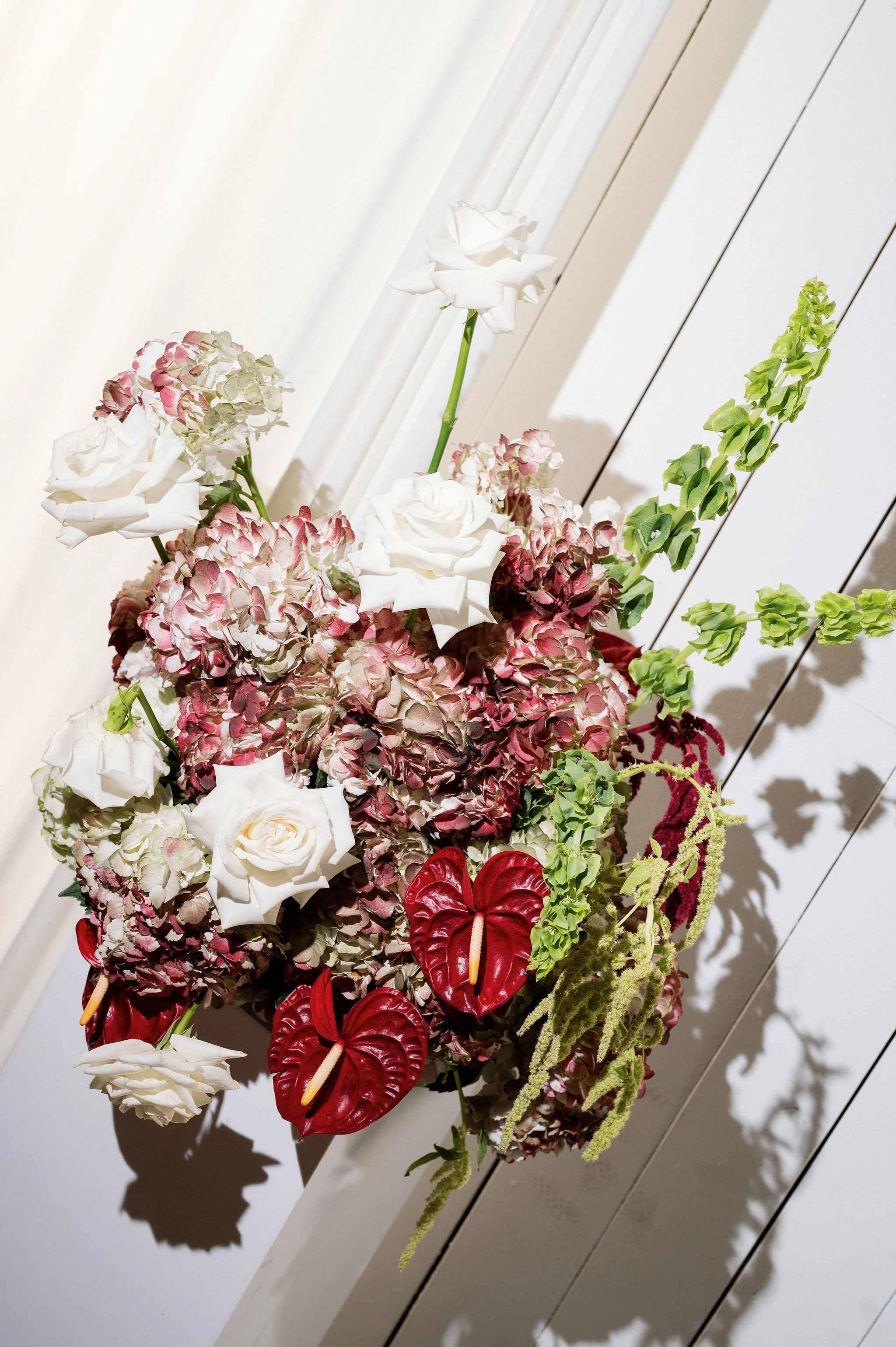 A floral arrangement with white roses, pink hydrangeas, red anthuriums, and green foliage, set against a white background with shadows.