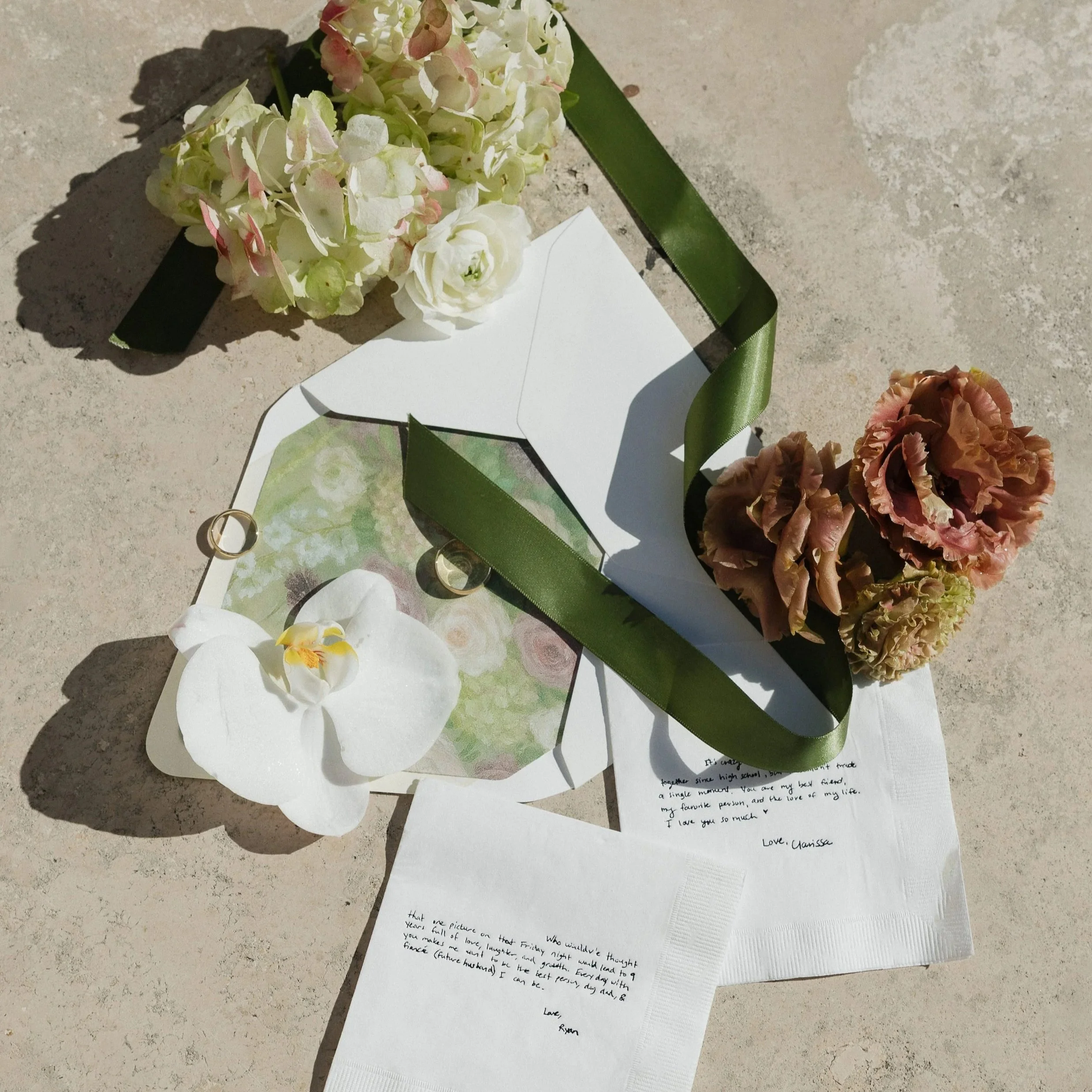 Flowers, letters, rings, and a white orchid on a concrete surface.