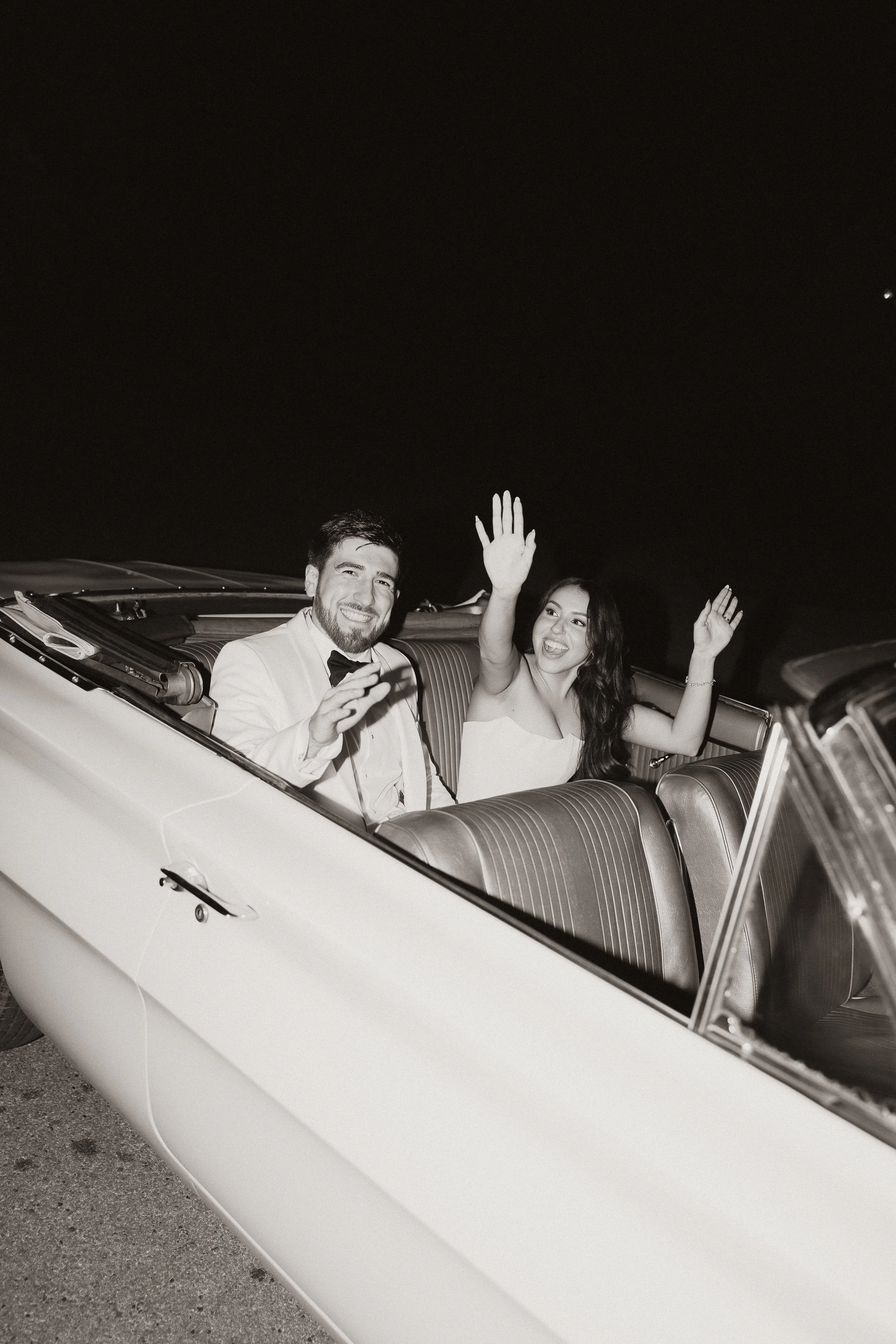 A bride and groom in formal attire sitting in a vintage convertible car at night, waving and smiling as they exit their wedding.