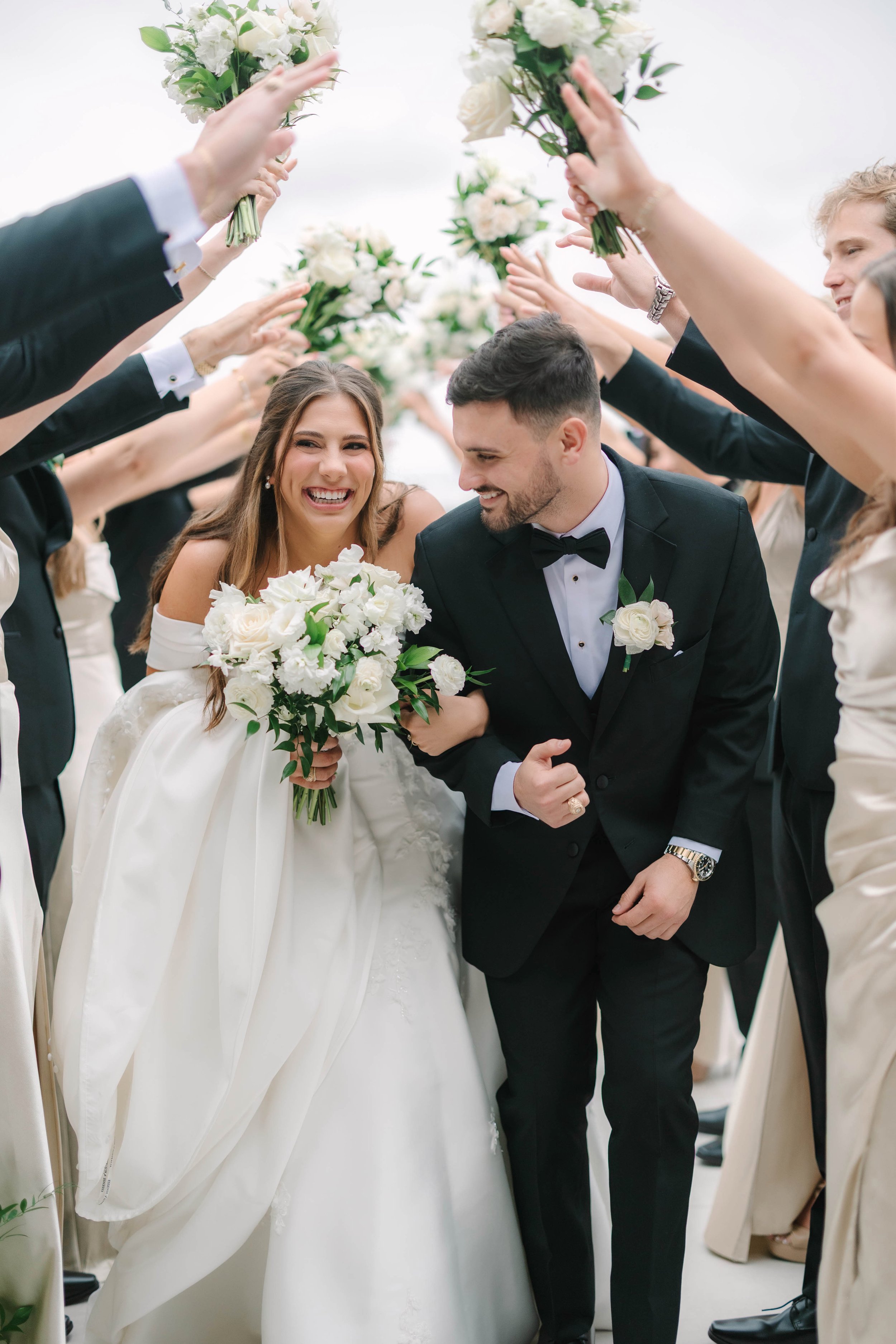 A bride and groom walking through a tunnel of raised hands and floral bouquets at their wedding, smiling and laughing.