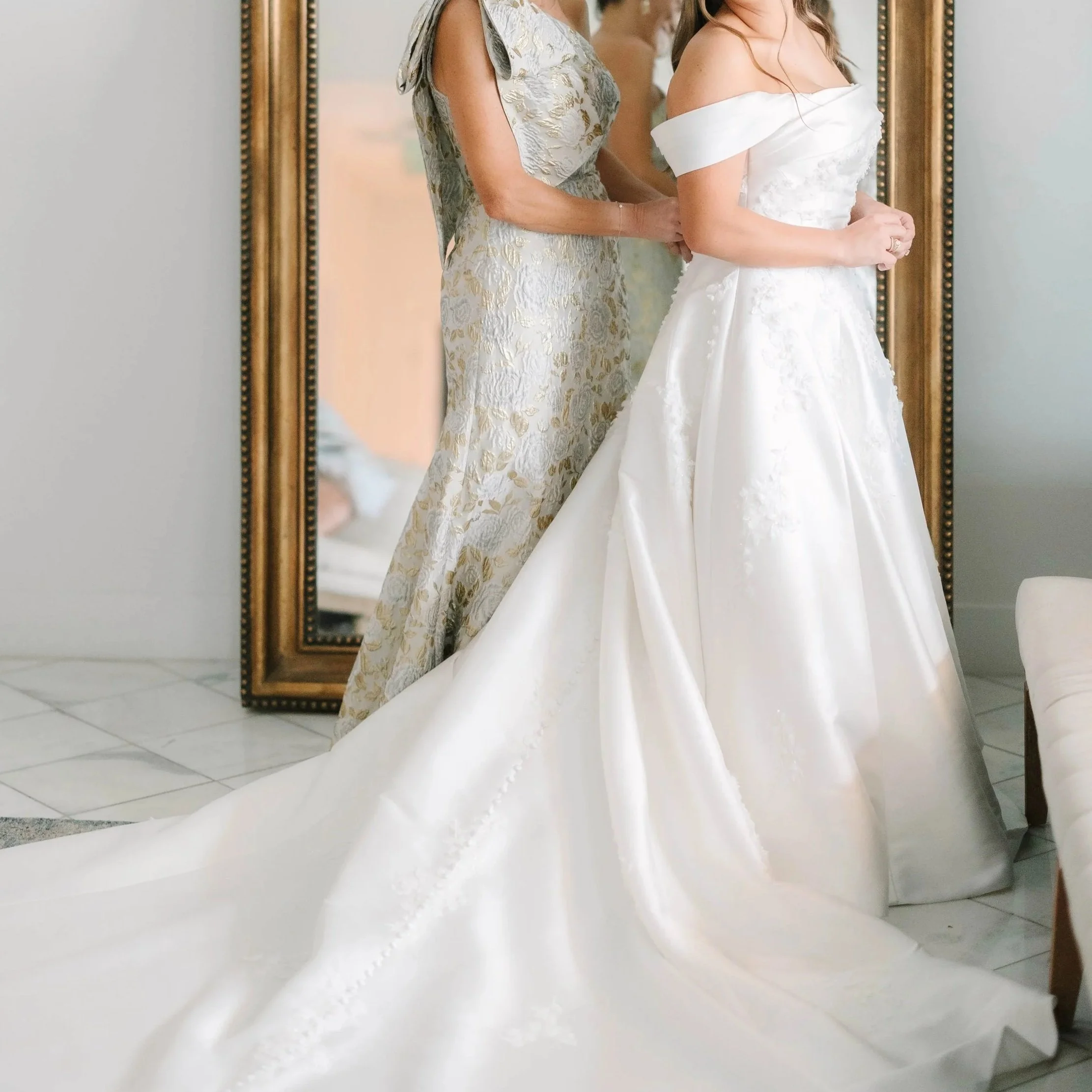 A bride in a white wedding gown with off-the-shoulder sleeves stands next to another woman in an elegant silver dress, with both reflected in a large mirror behind them.