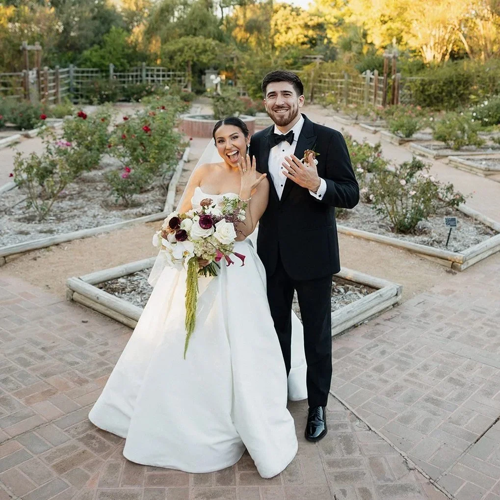 A bride and groom on their wedding day in a garden setting. The bride is wearing a strapless white wedding gown and holding a large bouquet of flowers. The groom is dressed in a black tuxedo with a bow tie. Both are smiling and showing off their wedding rings.