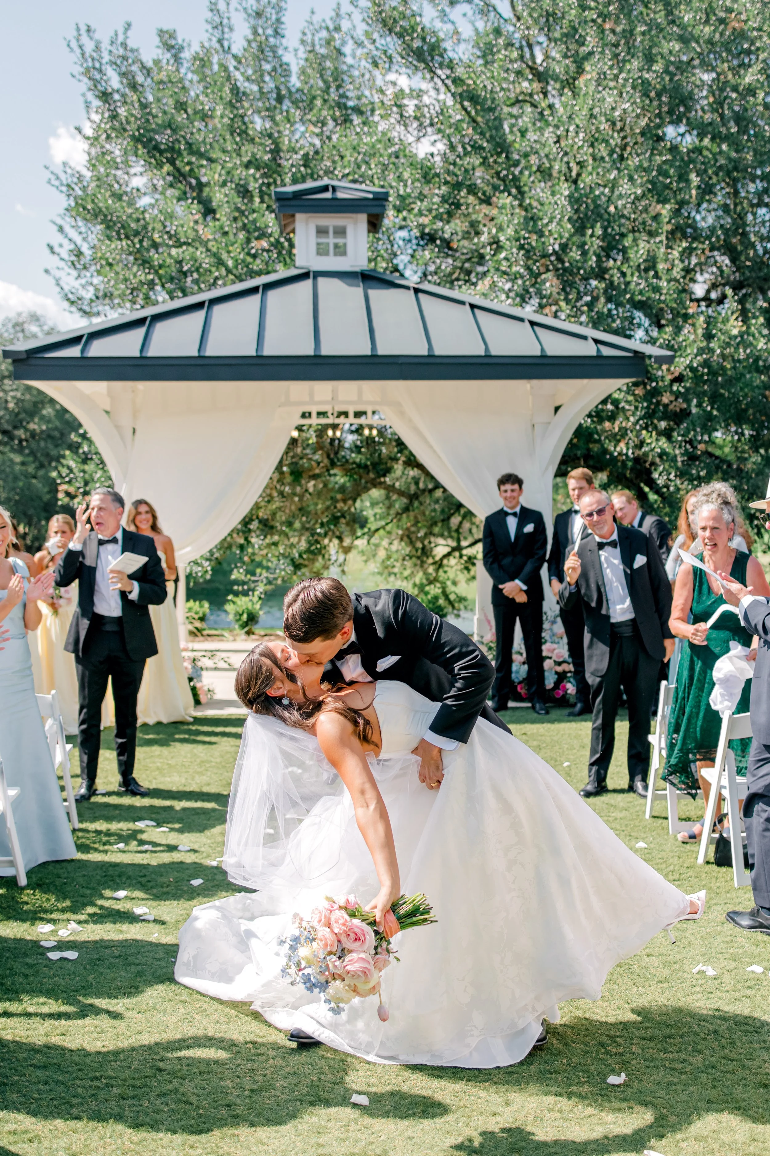 A bride and groom kiss during their outdoor wedding ceremony, surrounded by friends and family, under a gazebo and blooming trees.
