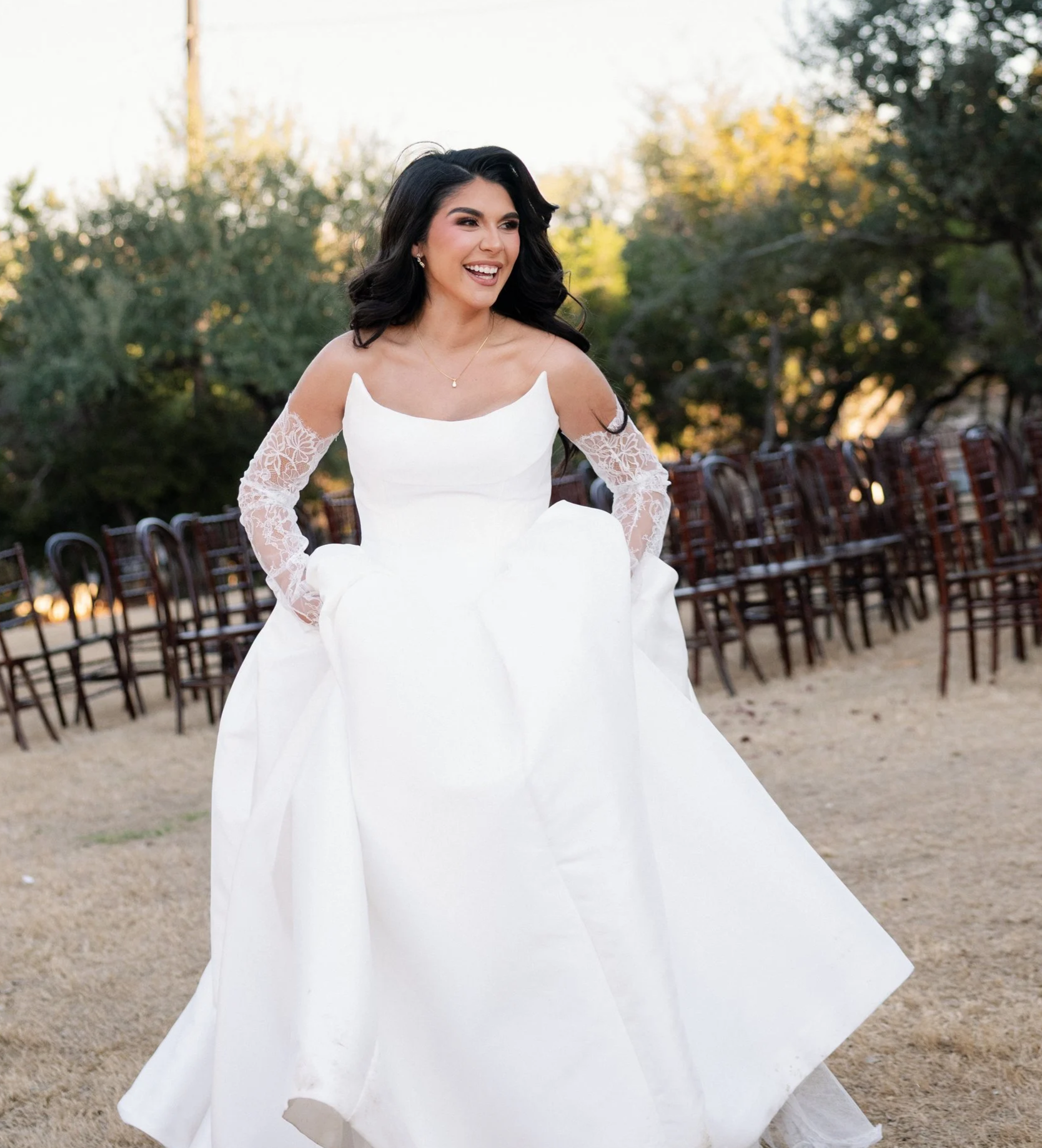 A woman in a white wedding dress with lace sleeves smiling outdoors with chairs in the background.