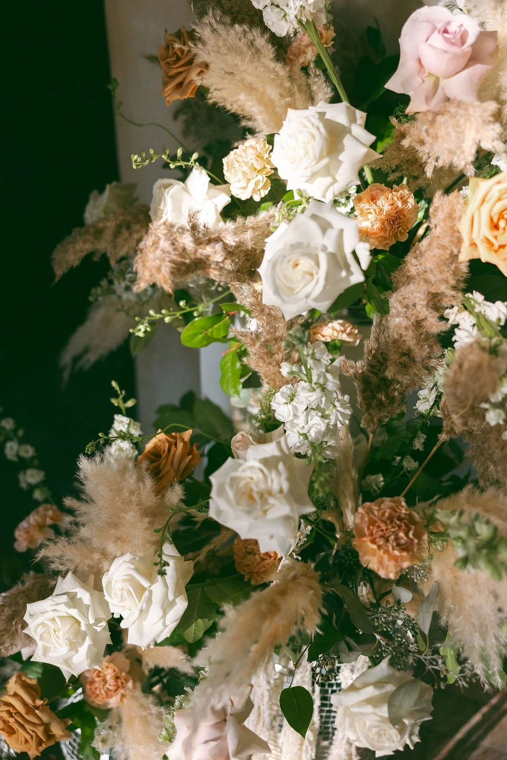 An arrangement of white, peach, and brown flowers, including roses, carnations, and pampas grass, with green foliage.