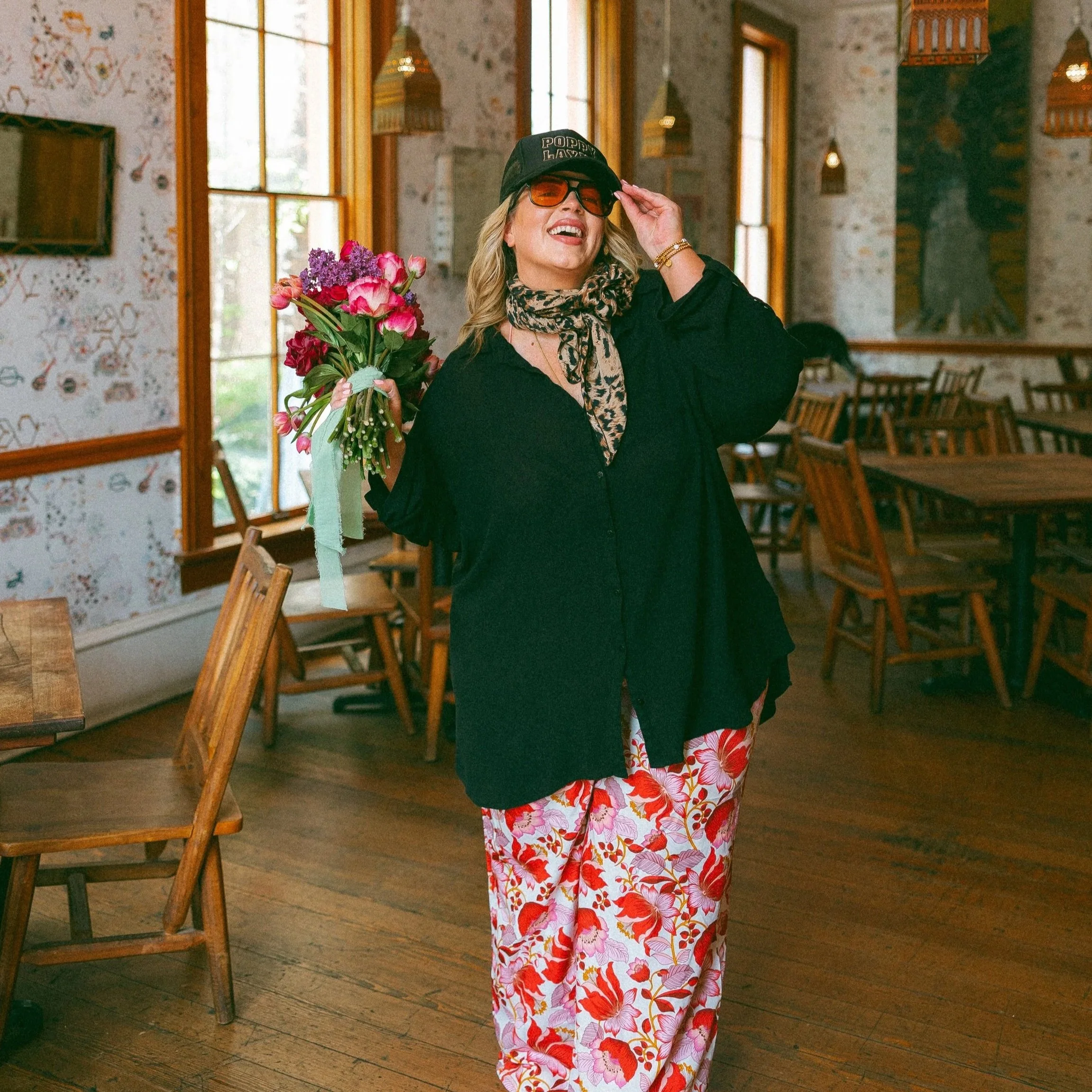 A woman holding a bouquet of flowers, wearing a black shirt, patterned pink and white pants, a leopard-print scarf, sunglasses, a baseball cap, and smiling inside a rustic cafe or restaurant.