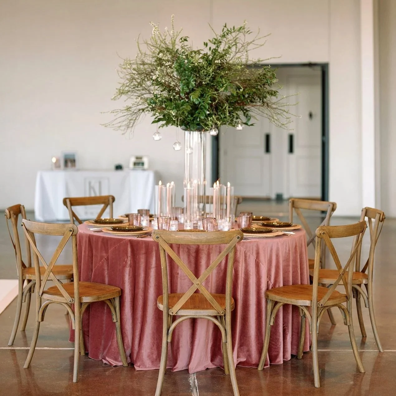 Round table with pink velvet tablecloth, wooden chairs, tall glass vase with a large floral arrangement, candles, and place settings in a decorated event space.