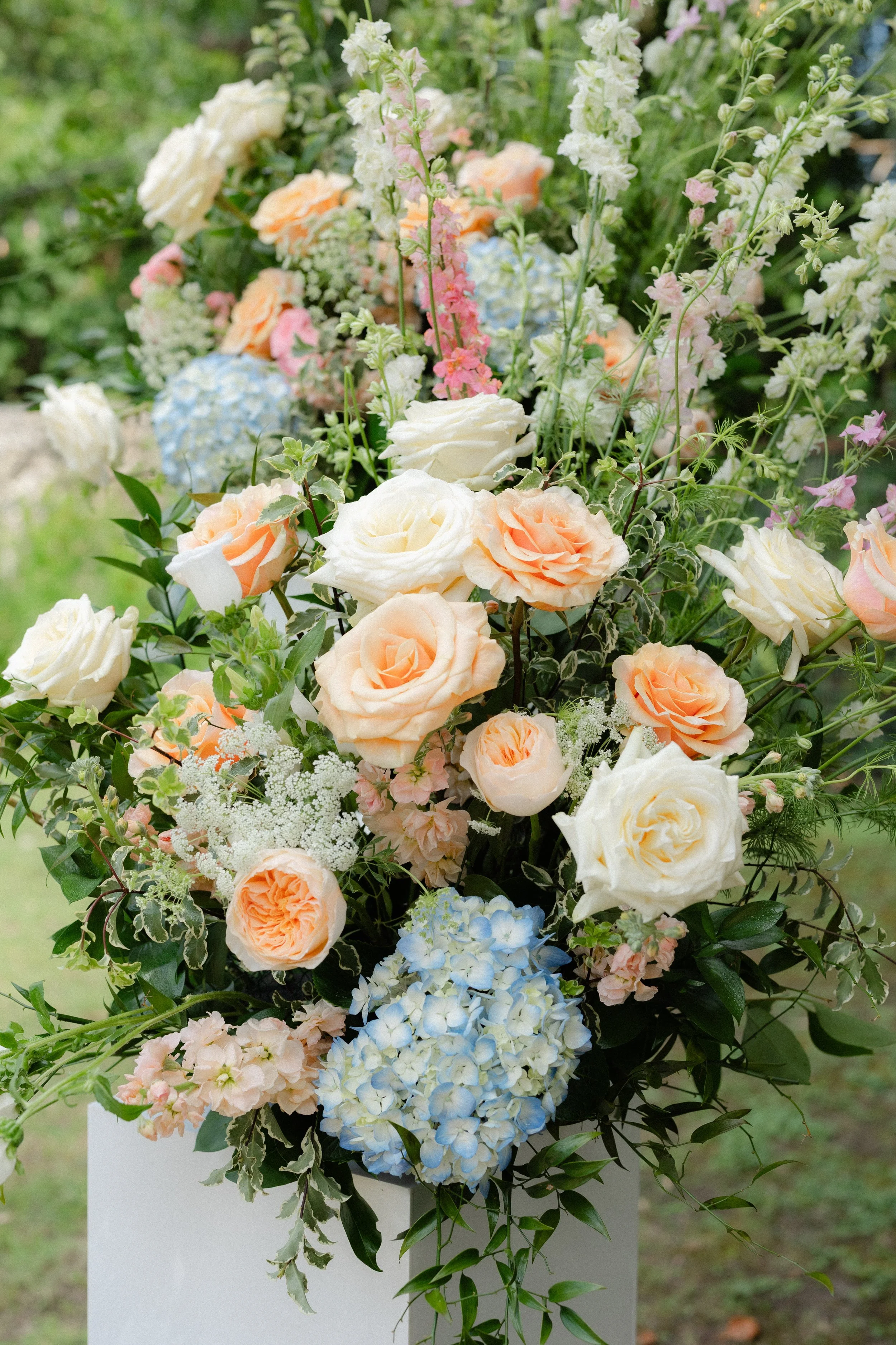 A floral arrangement with white, peach, pink, and blue flowers in a white vase outdoors.