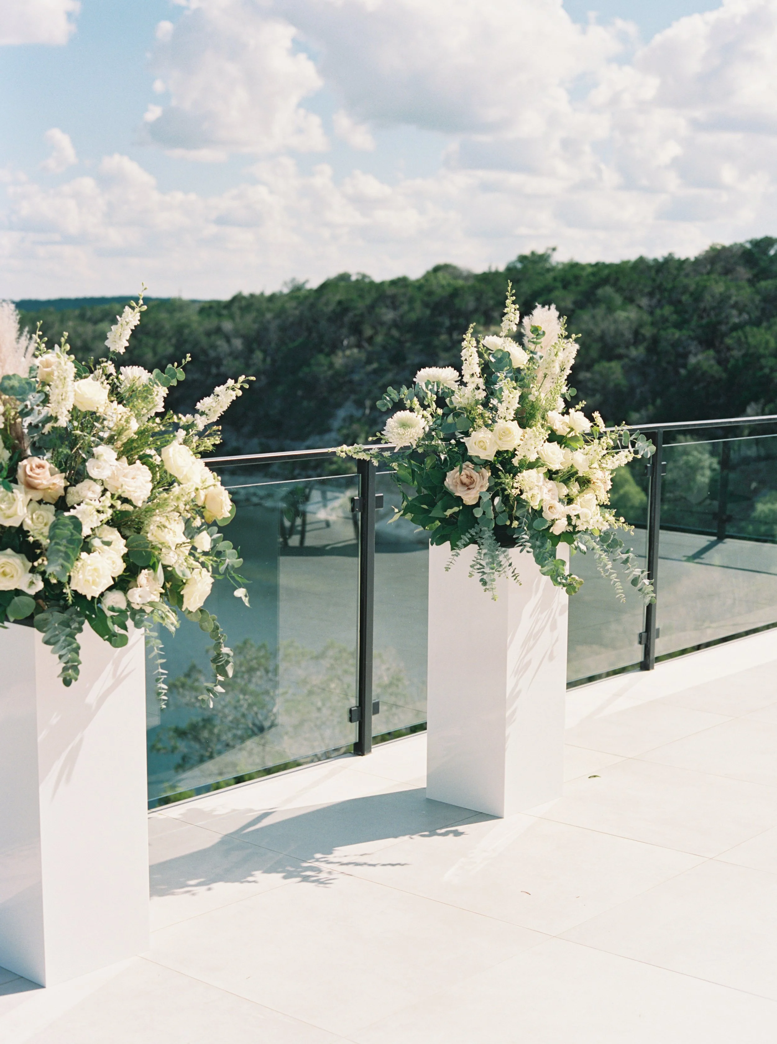 Two tall white flower arrangements on white pedestals on a balcony with glass railing, overlooking a green landscape and blue sky with clouds.