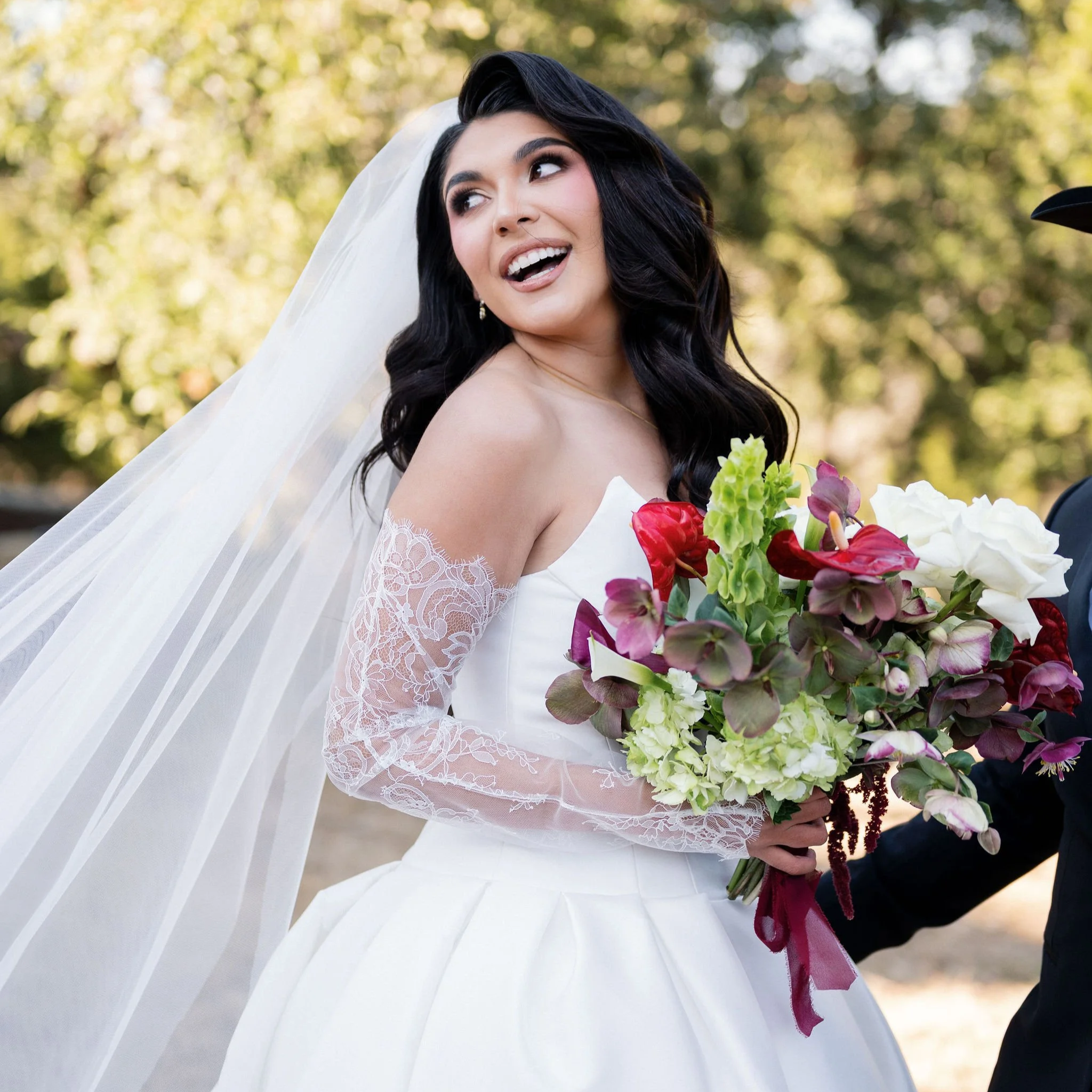 A bride in a white wedding dress holding a colorful bouquet of flowers outdoors, smiling and looking to her left.