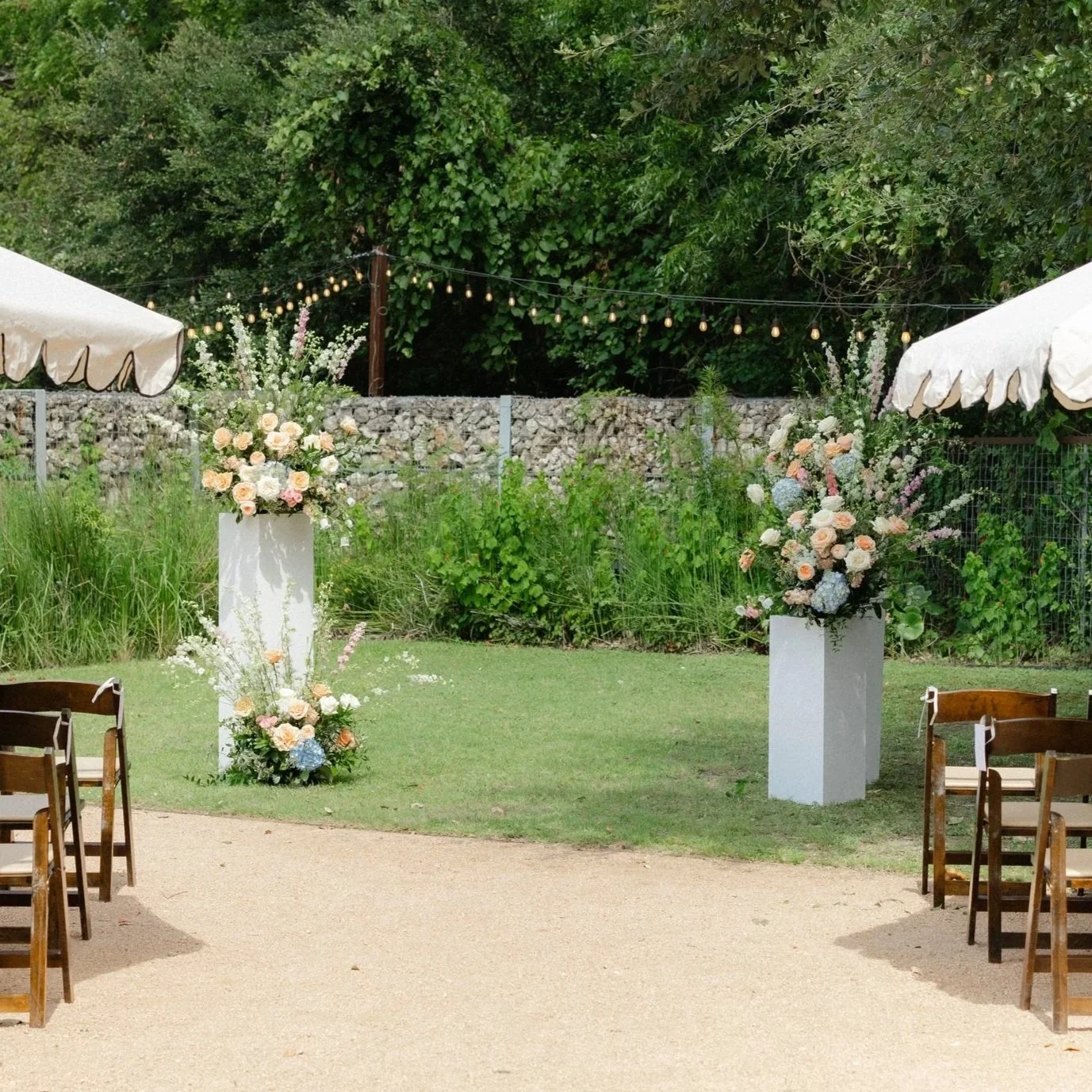 Outdoor wedding ceremony setup with floral arrangements on tall white pedestals and wooden chairs under a canopy, with string lights hanging in the background.