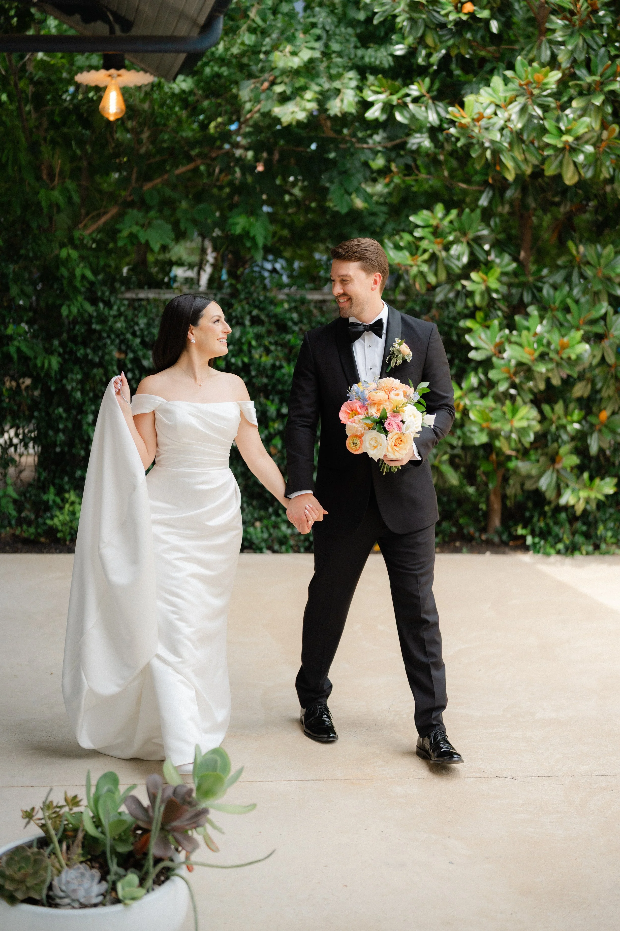 A newlywed couple walking hand in hand outdoors, with the groom holding a floral bouquet, surrounded by greenery.
