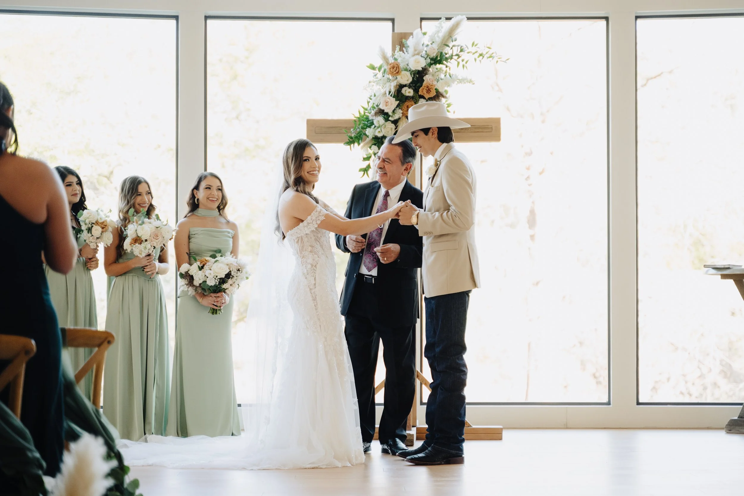 Bride and groom holding hands and smiling during wedding ceremony in front of near window with bridesmaids and bouquet.
