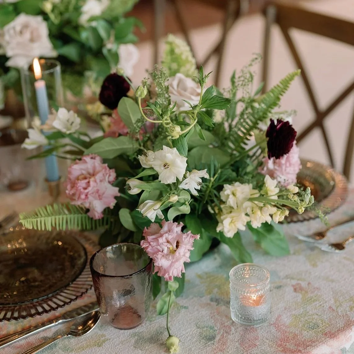 Floral centerpiece with pink, white, and dark red flowers on a decorated dining table with candles and glassware.