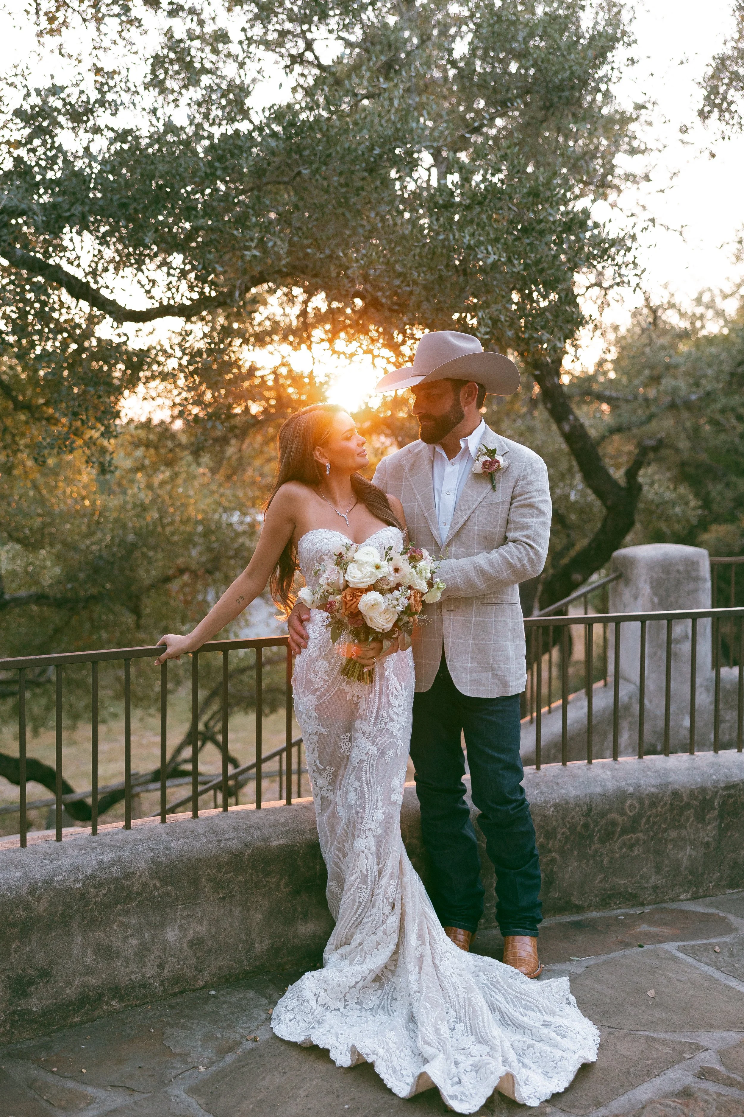 A bride and groom standing outdoors at sunset, with trees in the background. The bride is wearing a white lace wedding gown and holding a bouquet, while the groom is wearing a light-colored plaid blazer, white dress shirt, dark jeans, and a cowboy ha