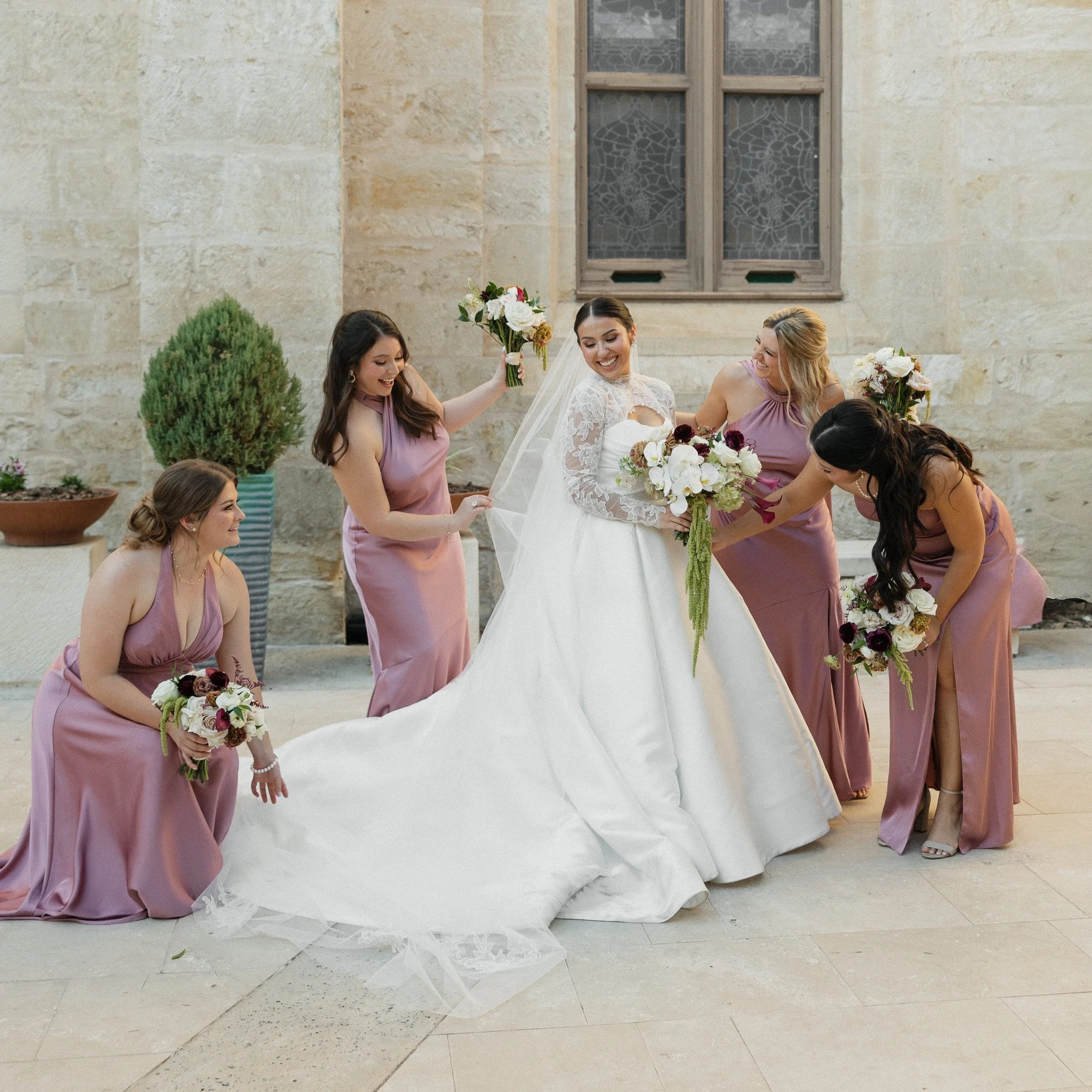 Bride in white wedding dress and veil with five bridesmaids in pink dresses holding bouquets, smiling and celebrating outside before a wedding ceremony.