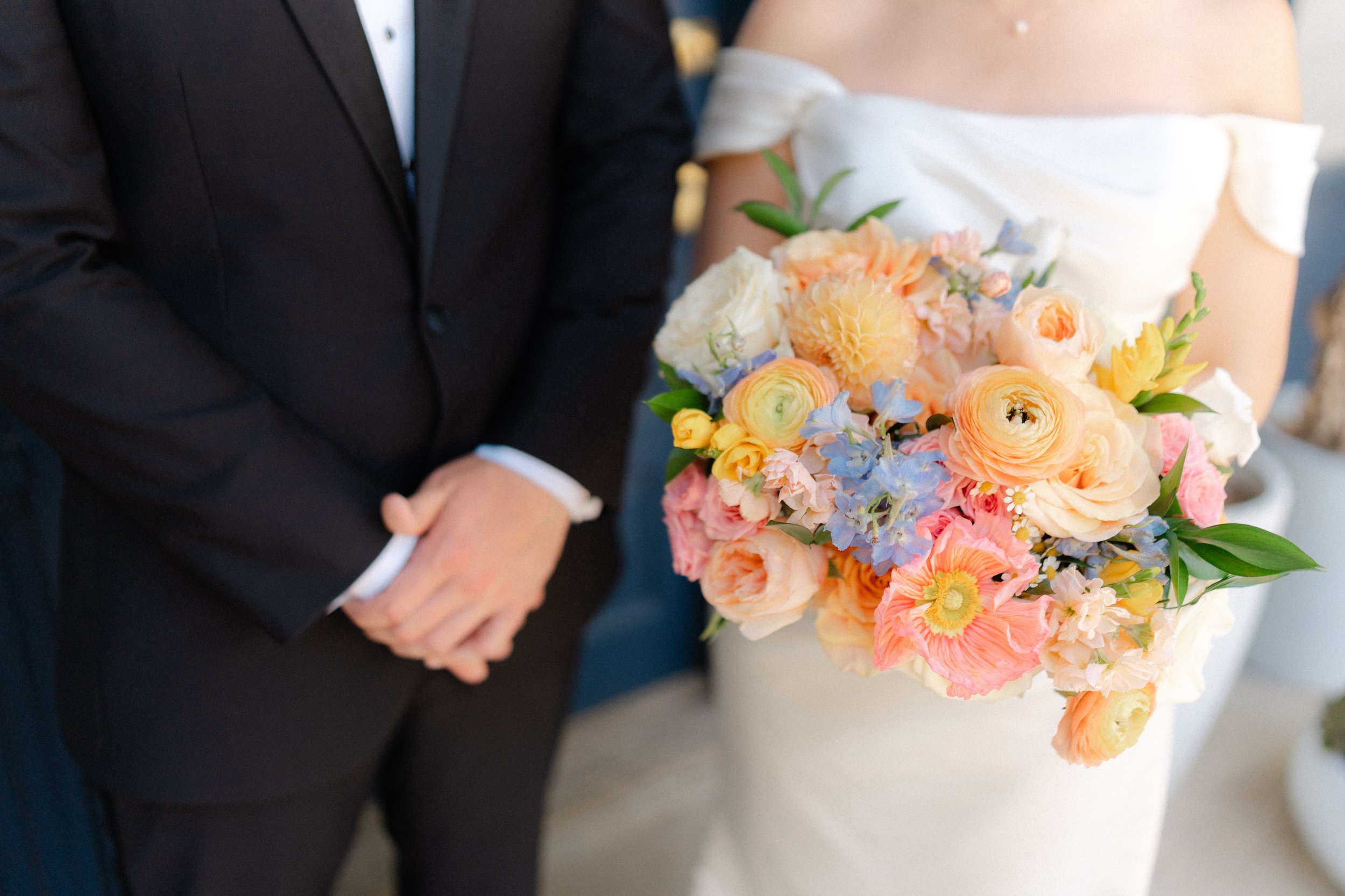 Close-up of a bride holding a pastel-colored bouquet of flowers and a groom standing next to her with folded hands at a wedding.