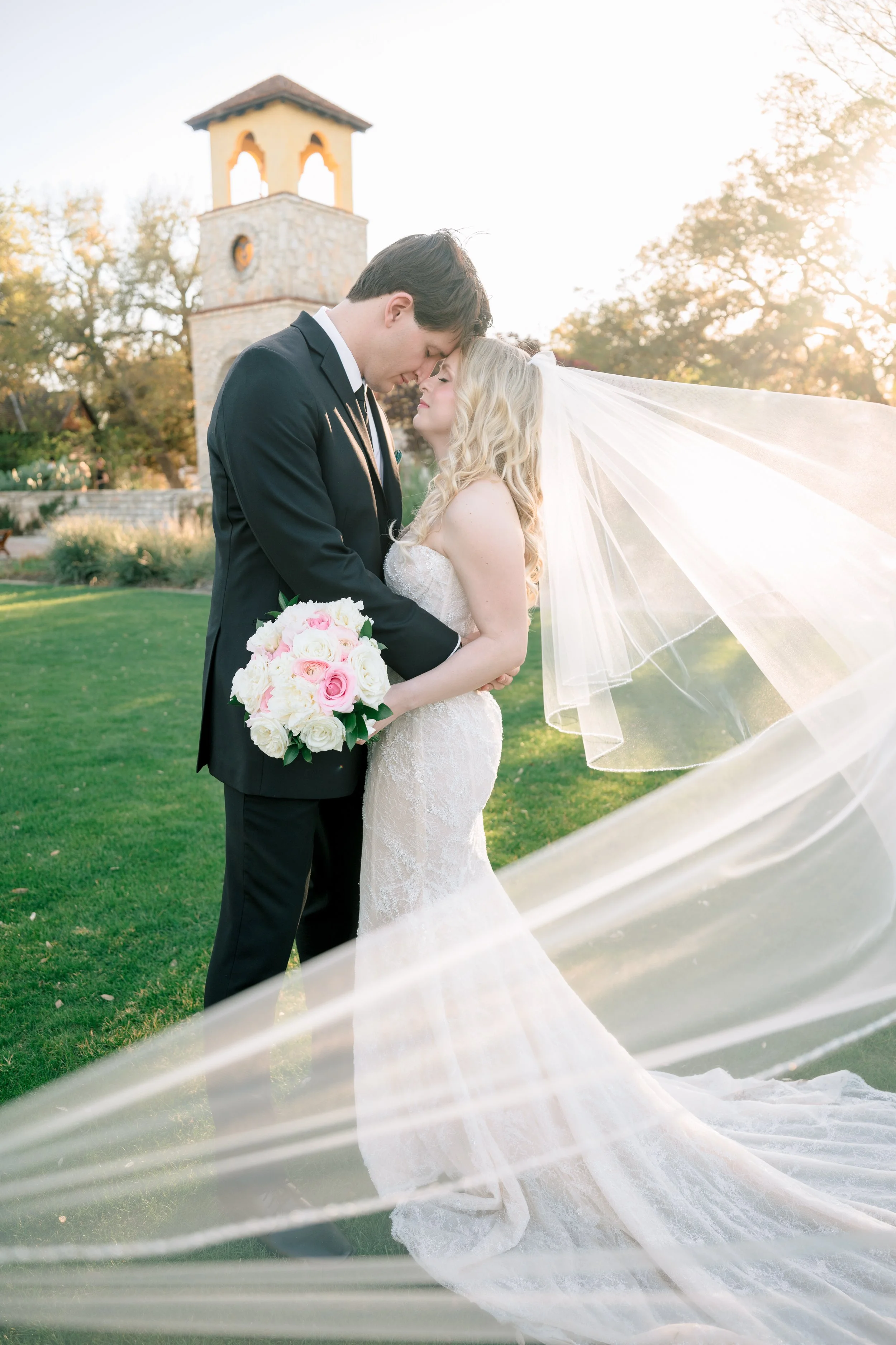 Bride and groom sharing a romantic moment outdoors on their wedding day, with the bride holding a bouquet of pink and white roses, and sunlight shining through trees.