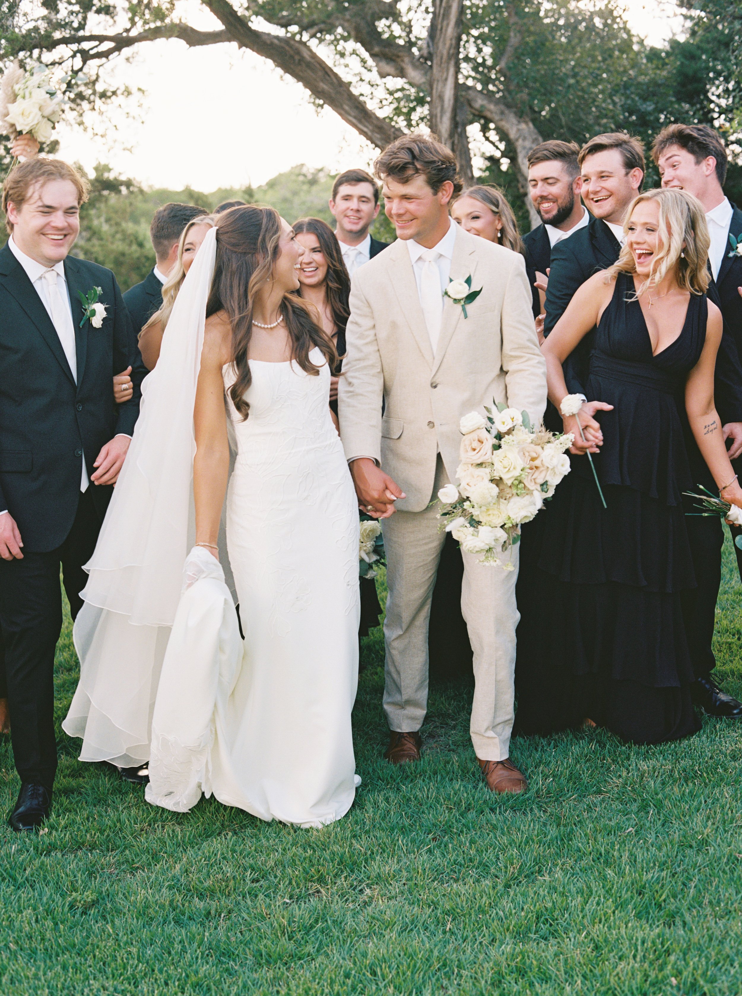 Wedding party outside with the bride, groom, and multiple guests smiling and holding hands, with a large tree and greenery in background.