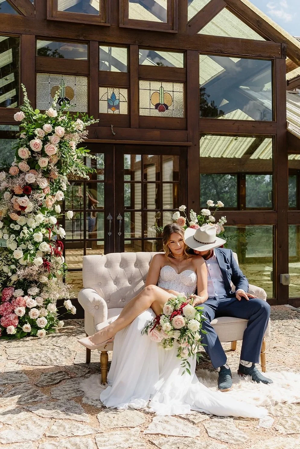 A bride and groom sit on a vintage sofa outside a glass and wood greenhouse, surrounded by pink and white flowers, with stained glass windows visible behind them.