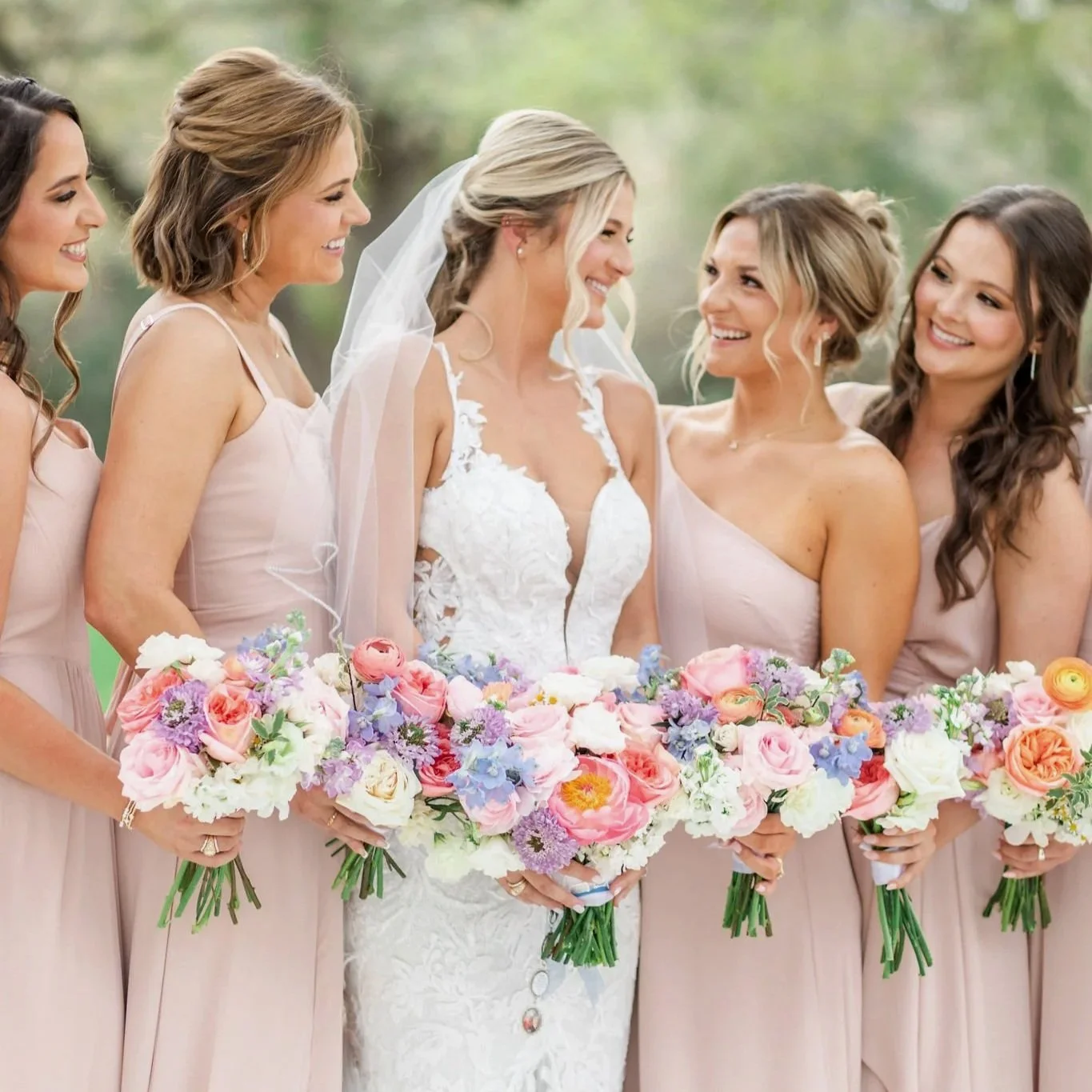 A bride in a white lace wedding dress and veil standing with five bridesmaids in blush pink dresses, all holding colorful bouquets of flowers, outdoors on a bright day.