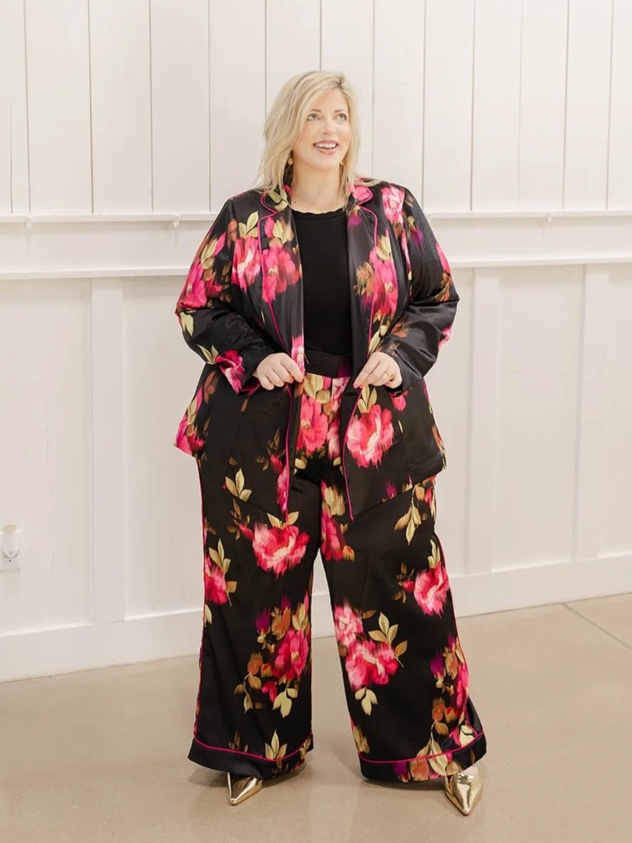 Woman wearing a black floral suit with pink and yellow flowers, standing in front of a white paneled wall.