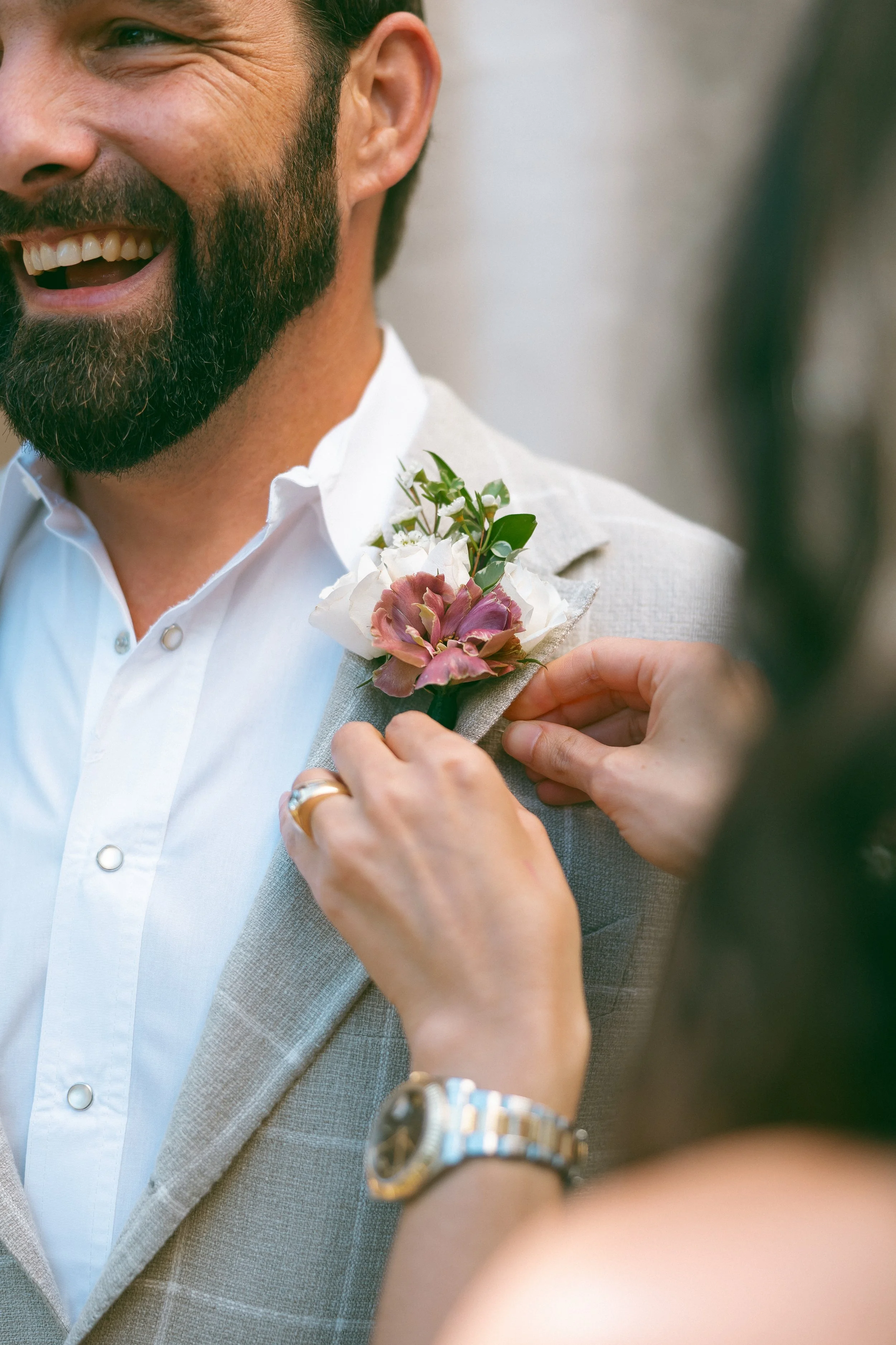 A person with a beard and smiling, wearing a white shirt and light gray suit jacket, has a woman pinning a pink and white boutonniere with greenery onto his lapel.