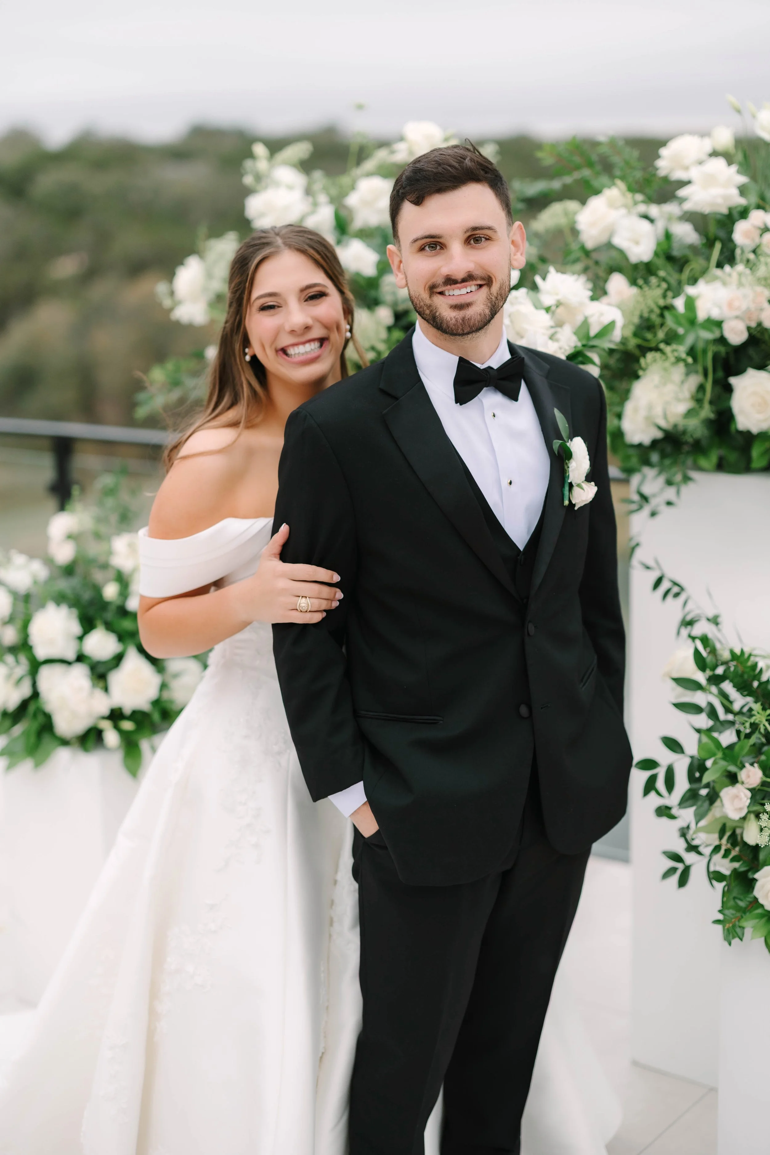 A bride and groom smiling at the camera during their wedding ceremony outdoors, surrounded by white floral arrangements.