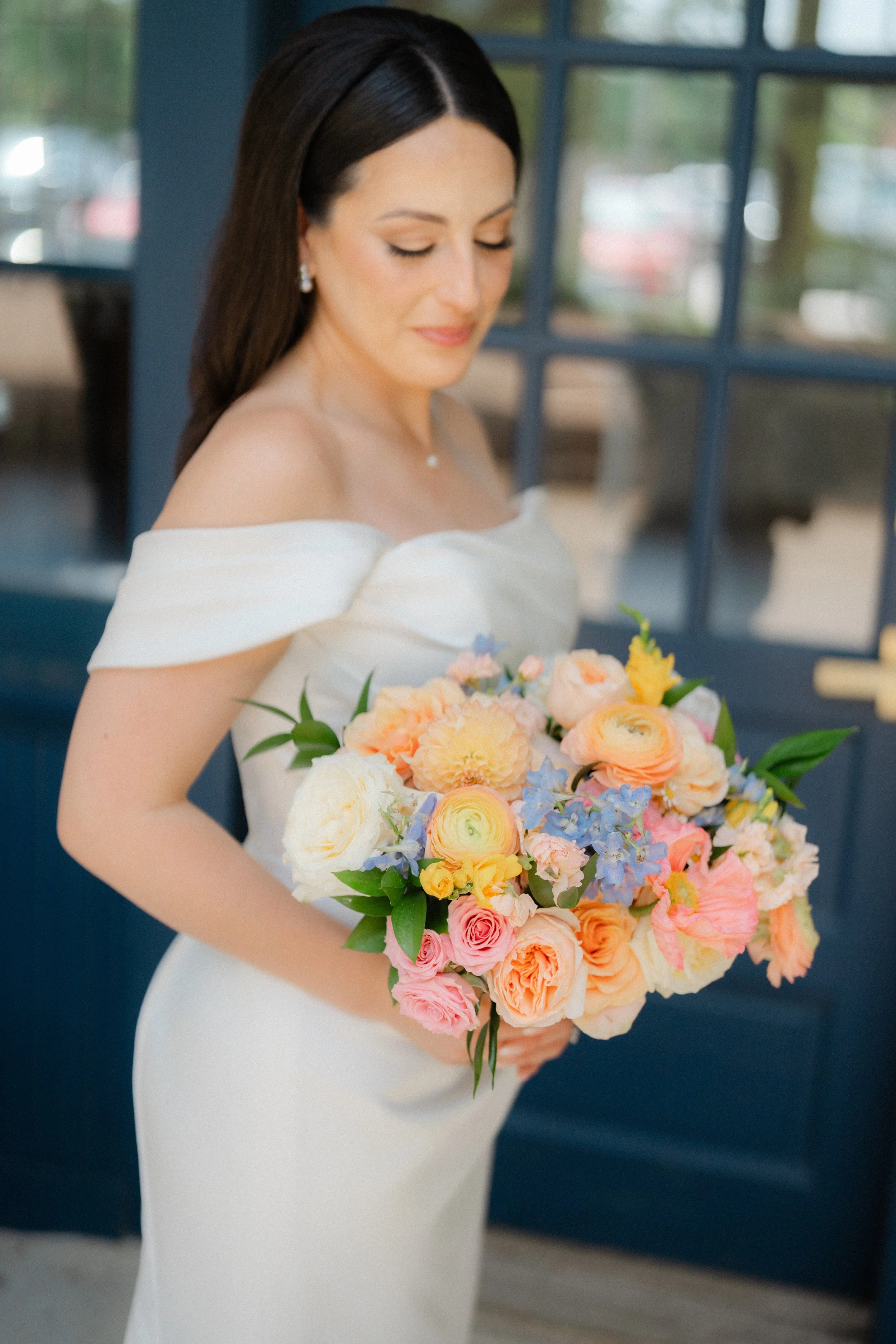 Bride holding a large colorful bouquet of flowers, wearing an off-the-shoulder white wedding gown, with a blue door and window in the background.