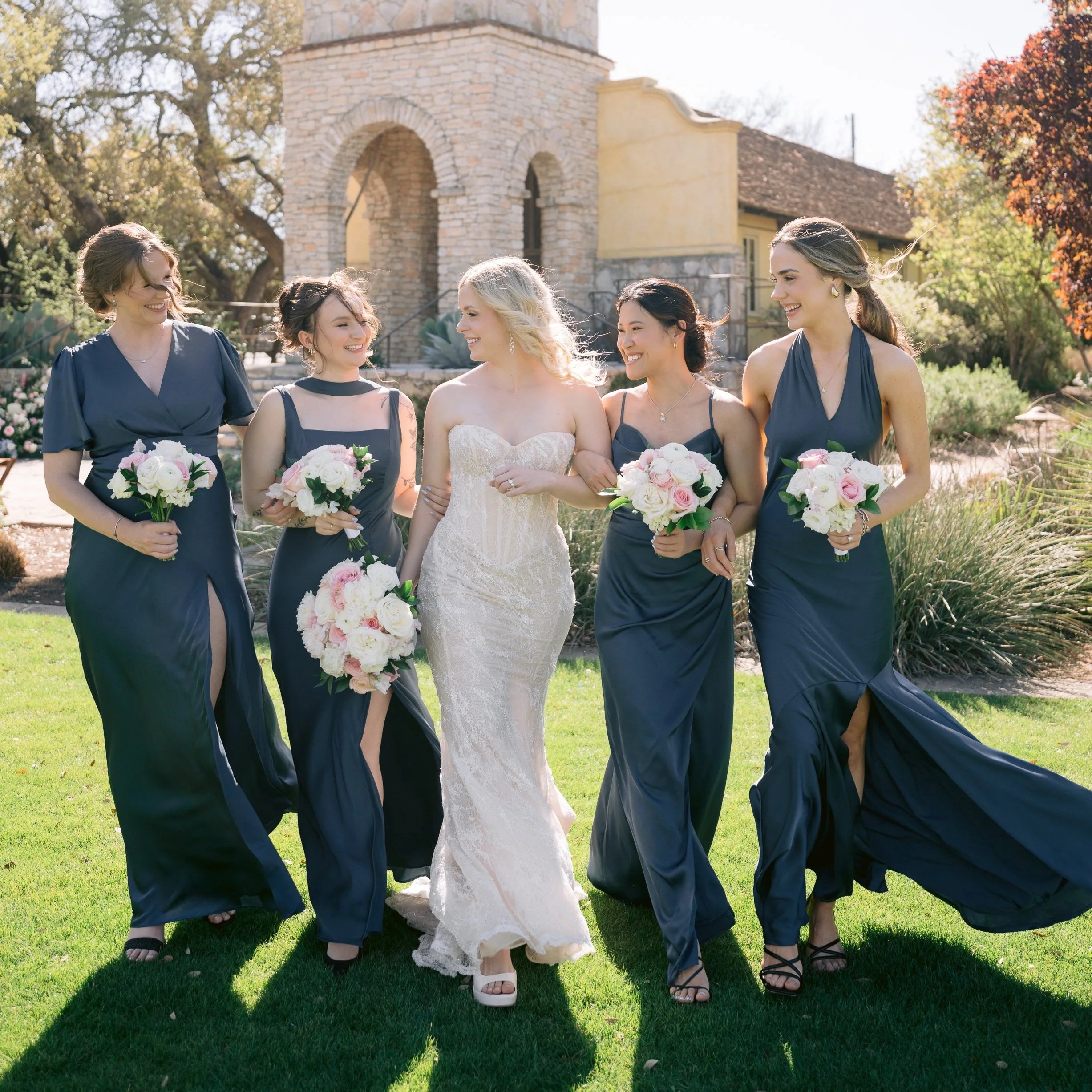 Bride in a white wedding dress walking with five bridesmaids in navy blue dresses, holding bouquets of pink and white flowers, outside on a sunny day near a stone building with trees and greenery in the background.