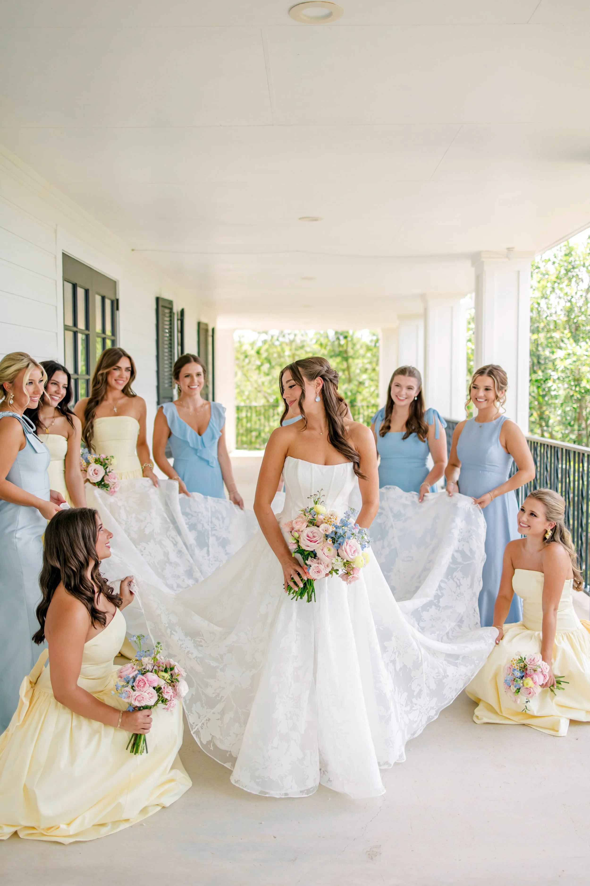 Bride in white wedding gown holding a bouquet, surrounded by her bridesmaids in pastel dresses, on a porch with greenery in the background.