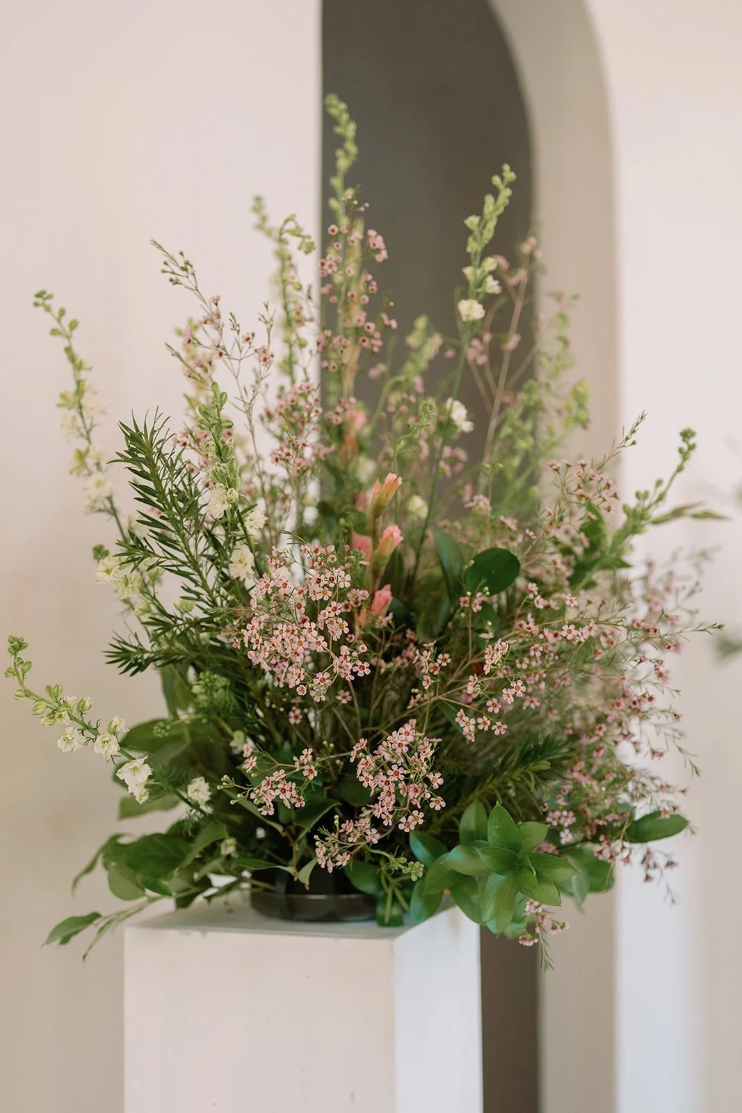 A bouquet of mixed flowers with pink, white, and green foliage in a black vase, placed on a white pedestal with a neutral background.