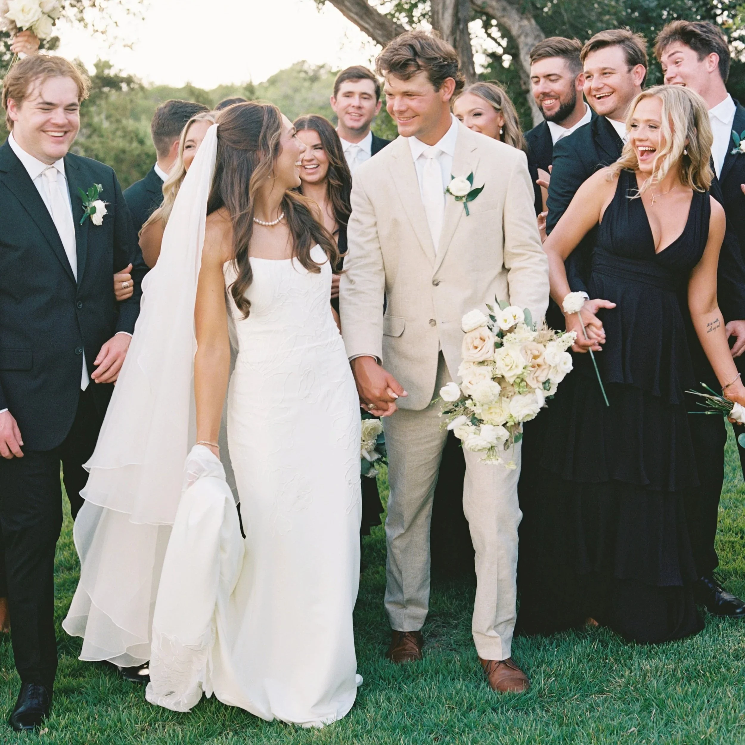 A wedding celebration outdoors with a bride and groom holding hands, surrounded by friends all smiling and laughing.