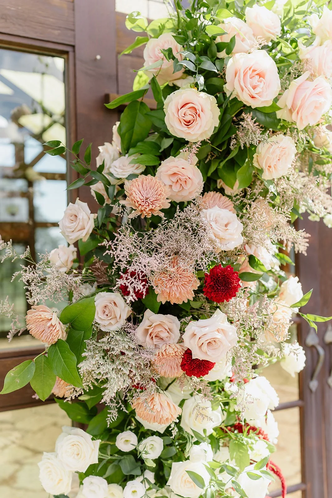 A cascading floral arrangement featuring light pink roses, peach carnations, white lisianthus, red dahlias, and delicate white filler flowers with greenery. The arrangement is set against a wood-paneled background with a window.