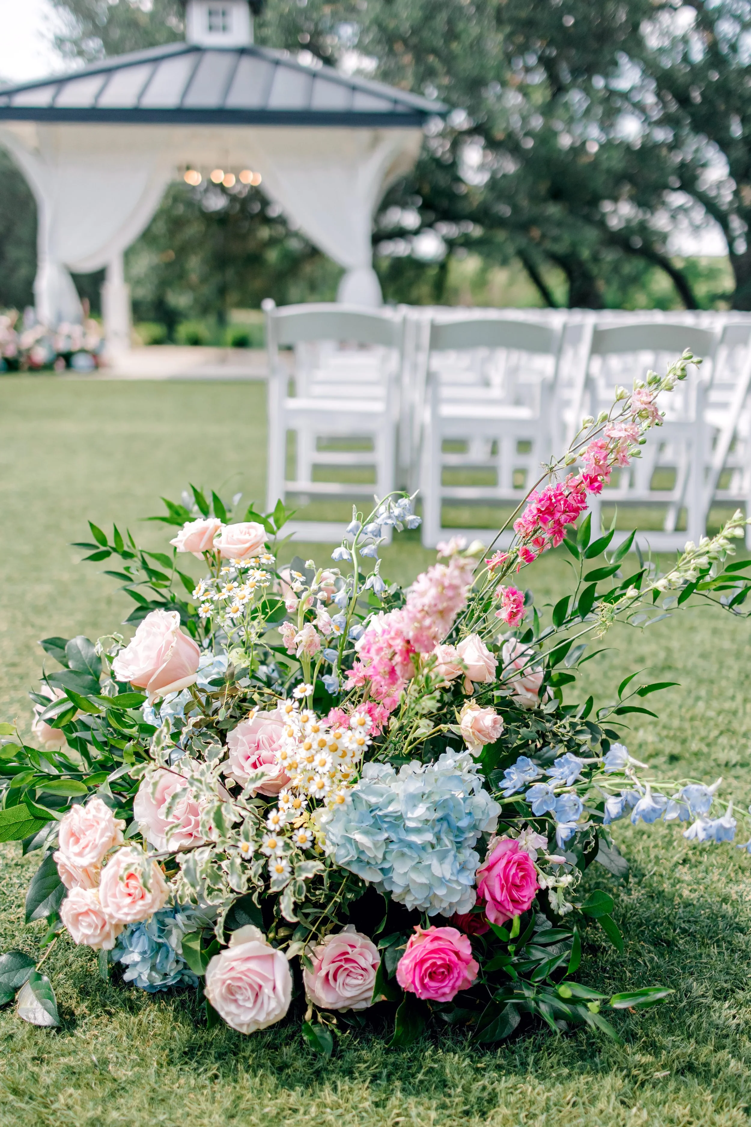 A colorful flower arrangement with pink, white, and blue flowers and green foliage on the grass near a white outdoor wedding setup with chairs and a gazebo in the background.