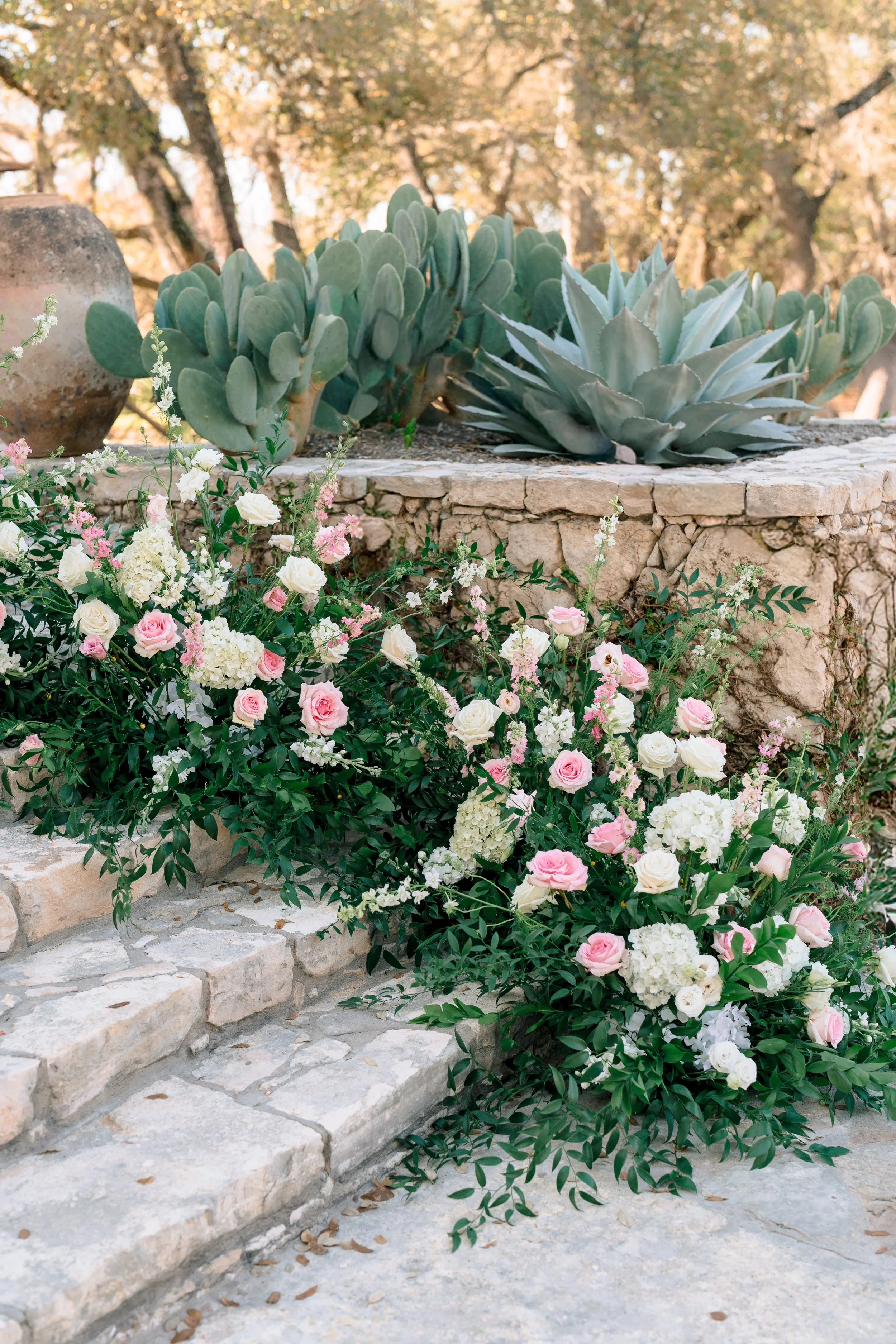 A floral arrangement with pink and white roses, white hydrangeas, and greenery on stone steps, with succulents and desert plants in the background.