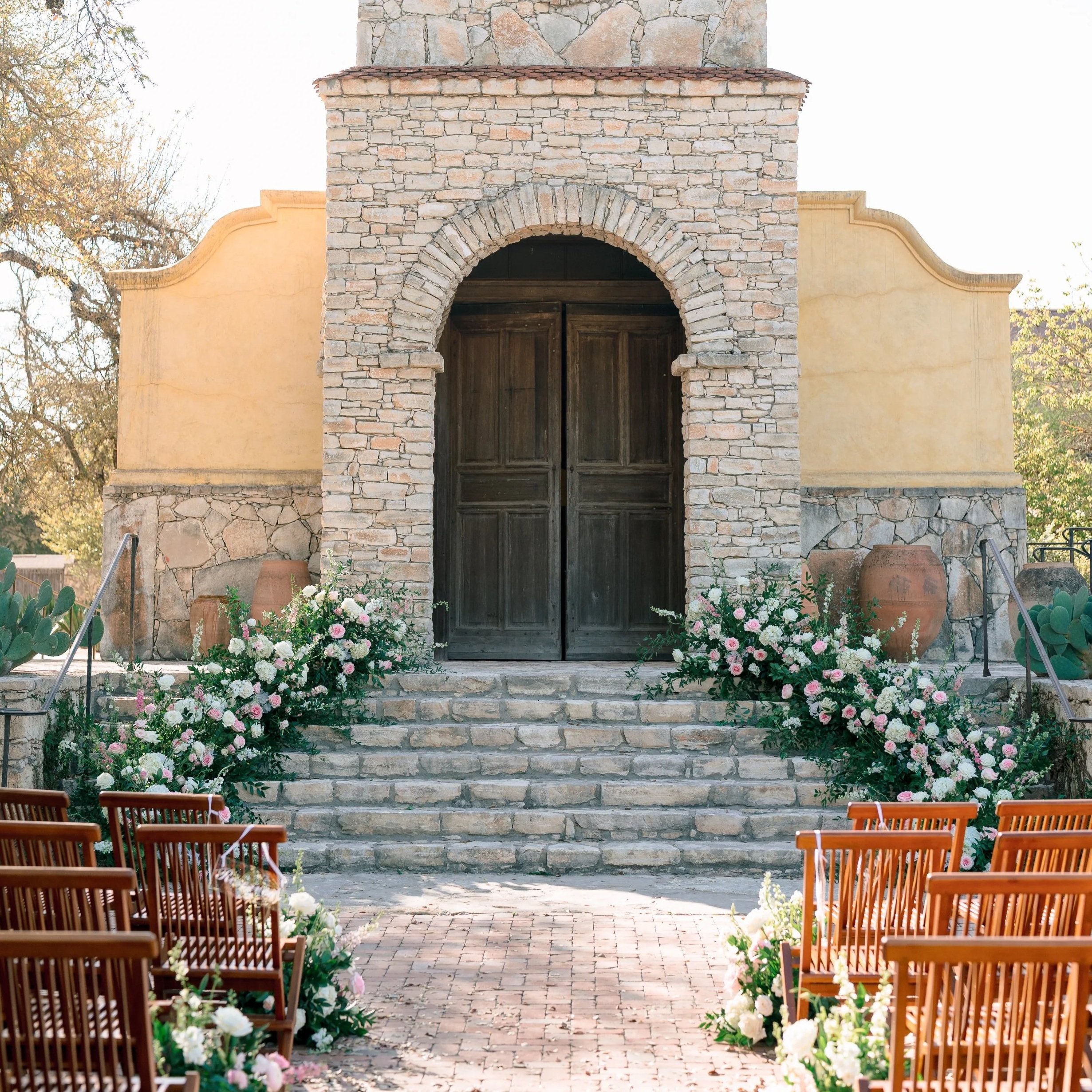 Outdoor wedding ceremony setup with wooden chairs and floral arrangements leading to stone steps and an arched wooden door in a rustic building.