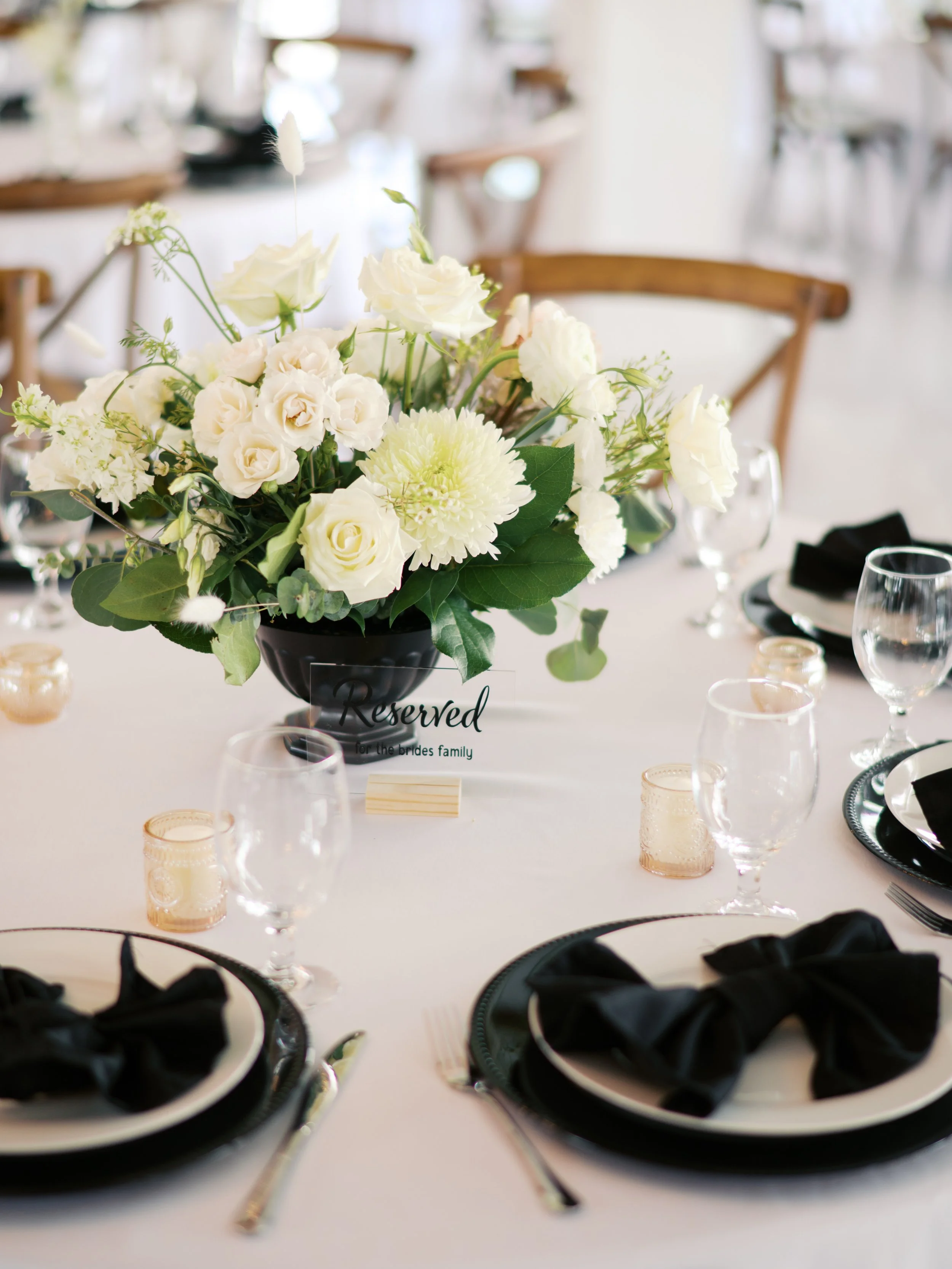 A decorated wedding reception table with a centerpiece of white flowers, surrounded by black and white dishware, glassware, black napkins, and small votive candles, with a 'Reserved' sign for the bride's family.
