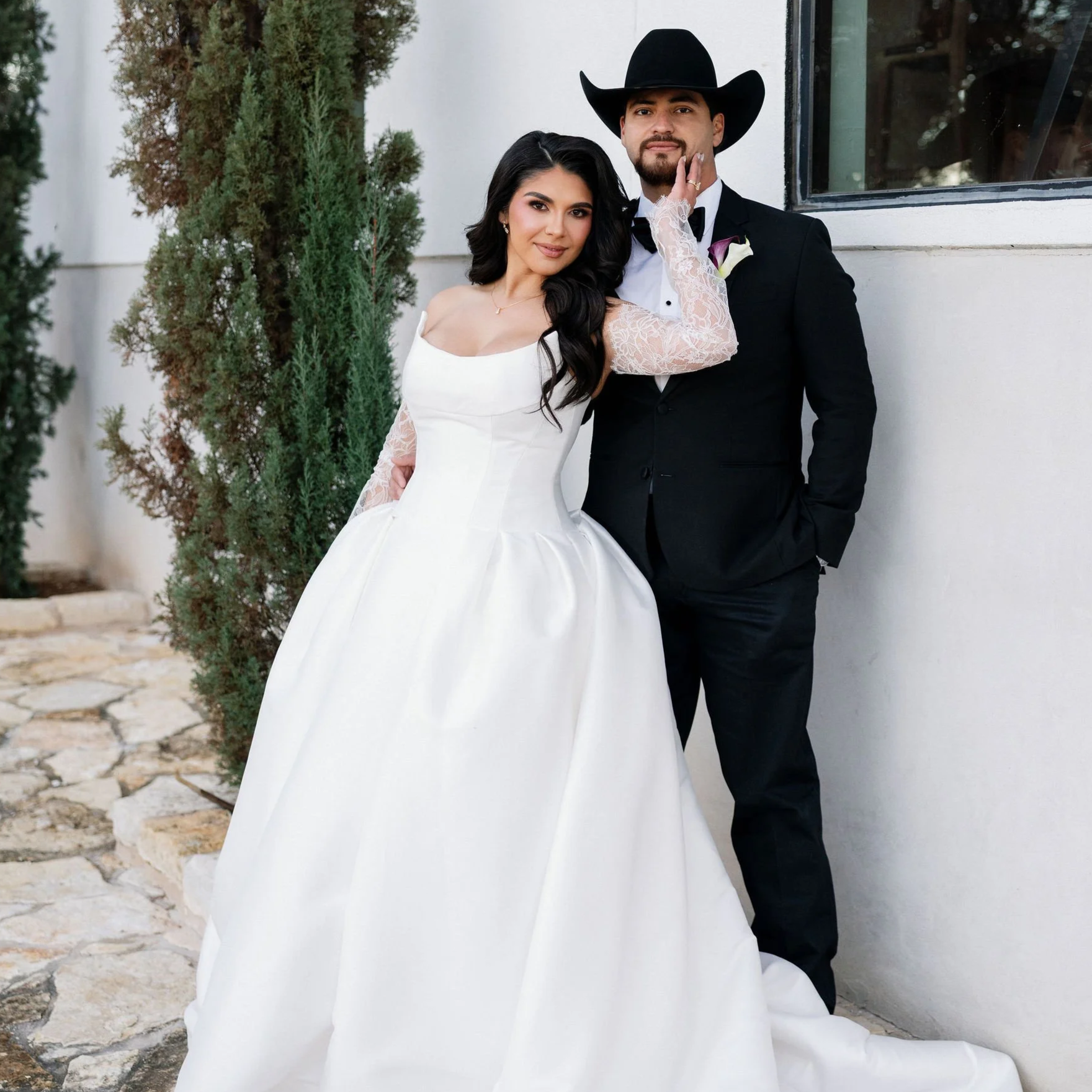 A bride in a white wedding dress and a groom in a black suit with a cowboy hat stand together outdoors, with trees and a white wall behind them.